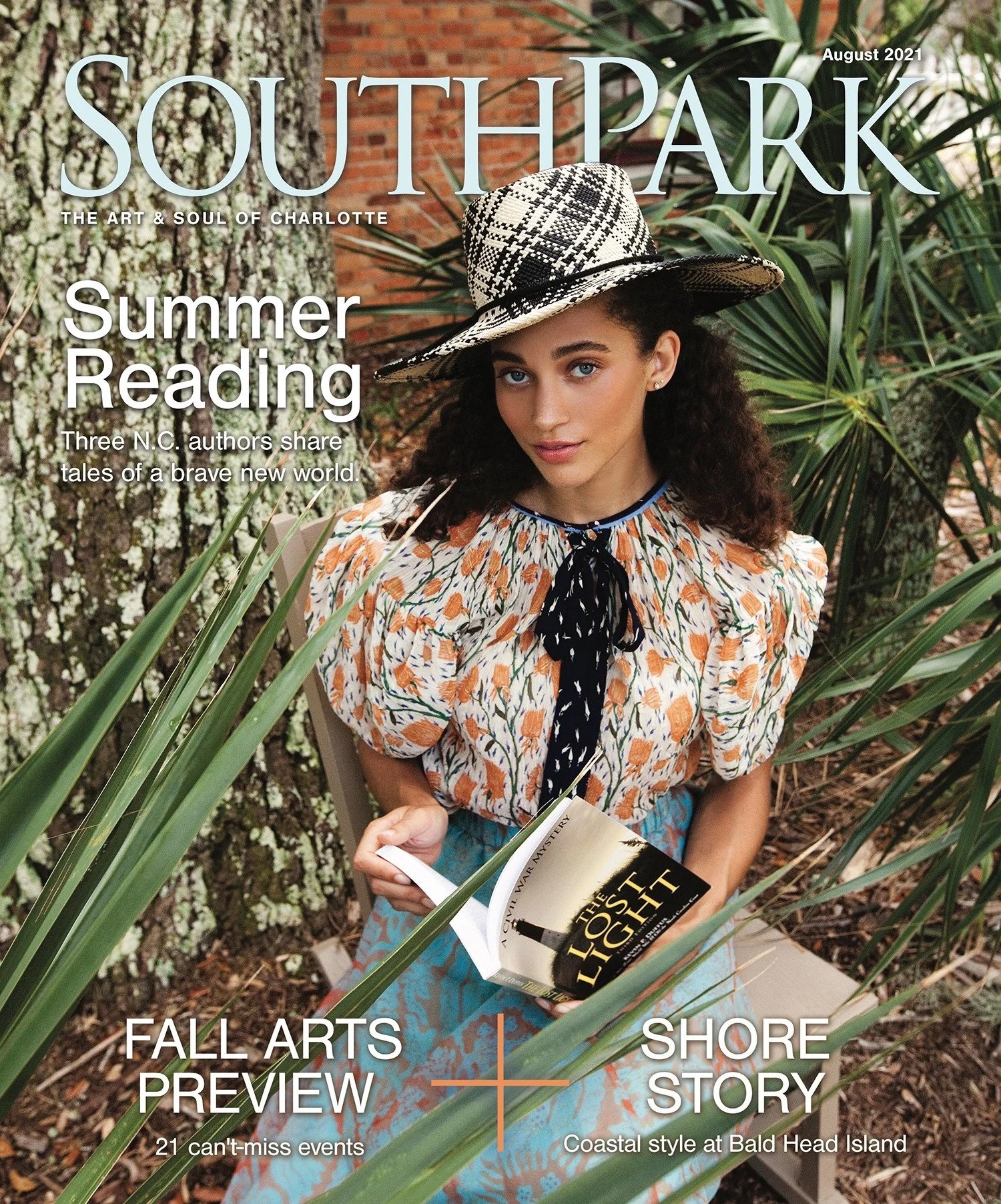 Young woman with curly hair wearing a hat and patterned blouse, holding a book outdoors among plants.