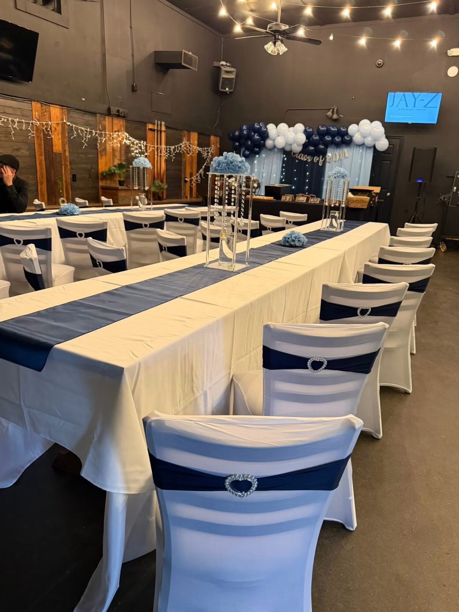 Event space with long tables, white chairs, blue table runners, and balloon decorations in blue and white, with string lights overhead.