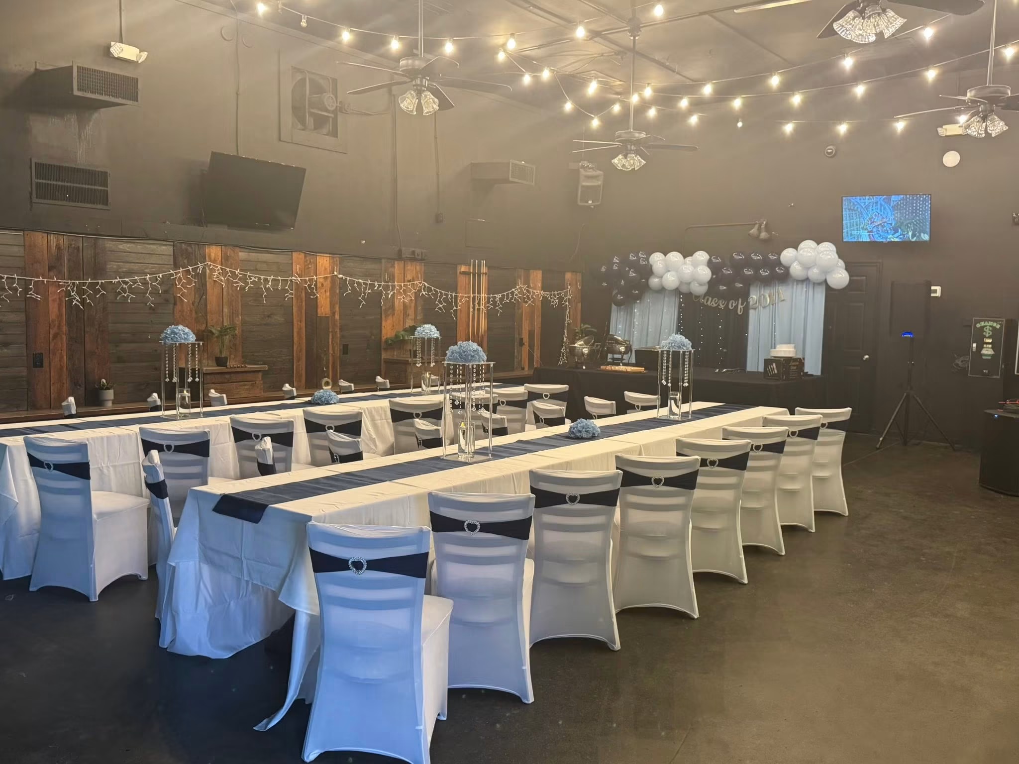 Event space with long tables, white chairs, and cloud decorations, illuminated by string lights and ceiling fans.