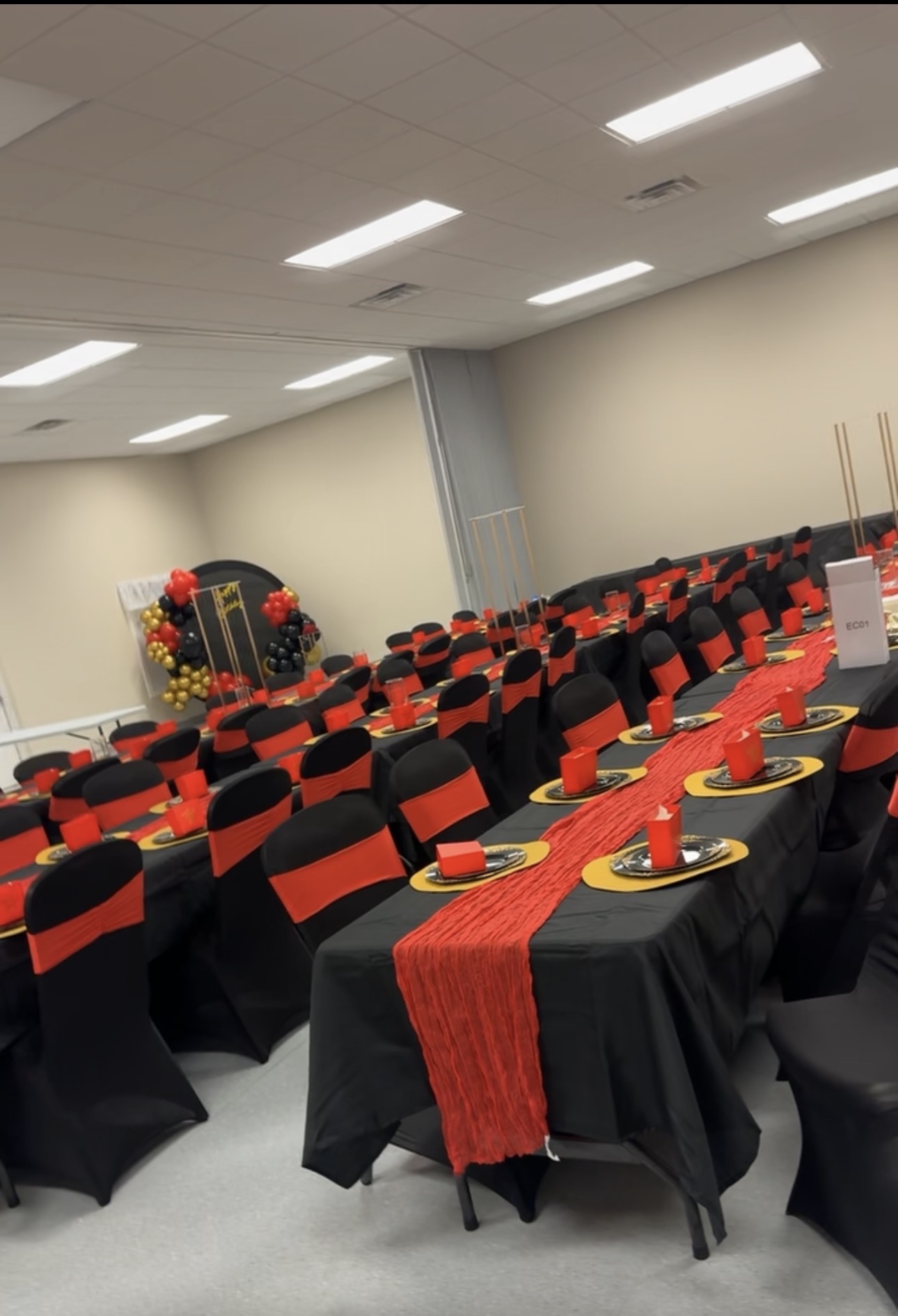 Banquet hall with rows of tables and chairs, decorated with red and black table settings and a stage with floral arrangements.