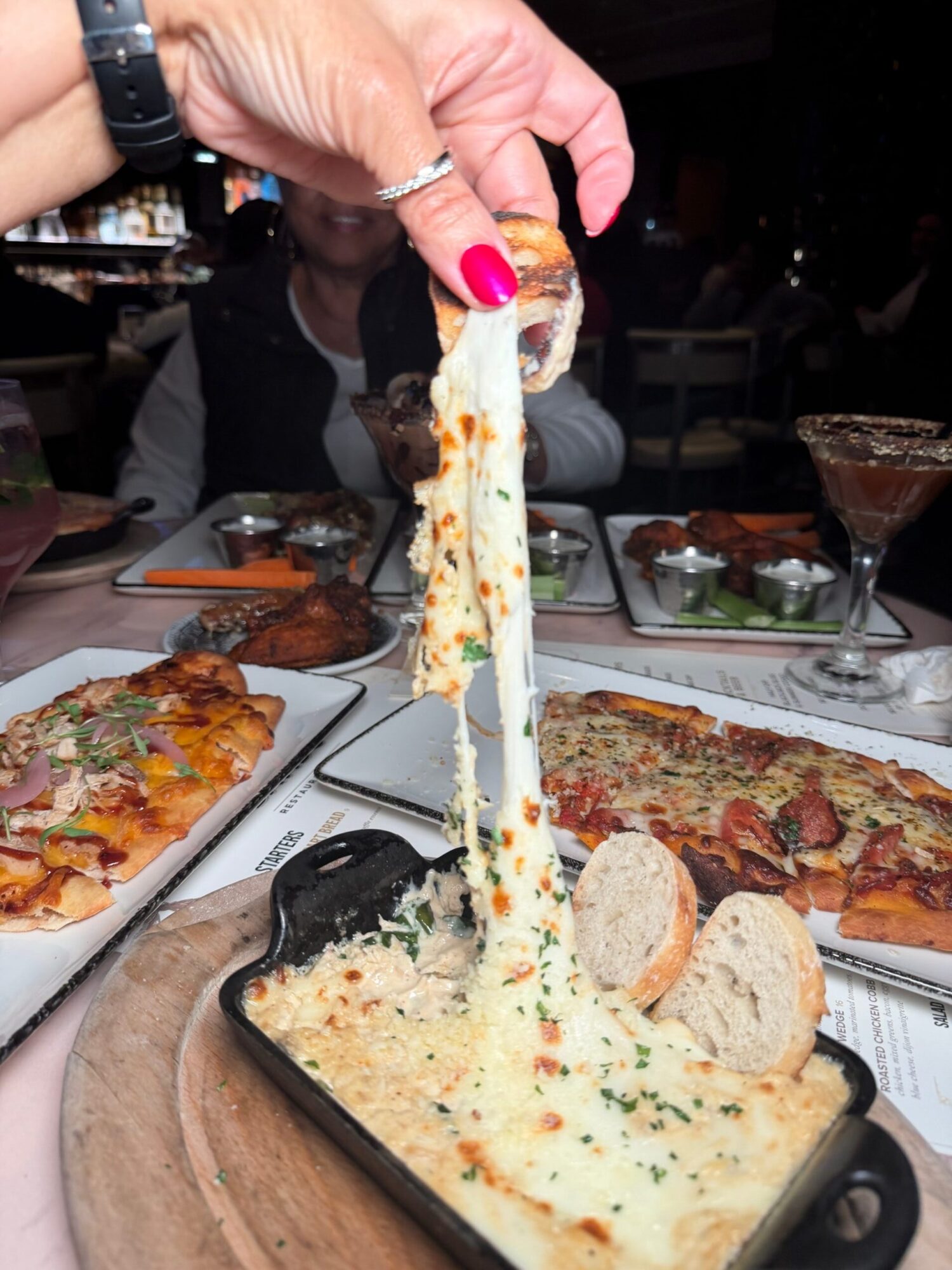 Person holding a slice of cheesy baked dish with bread, with pizza and other dishes on table in background.