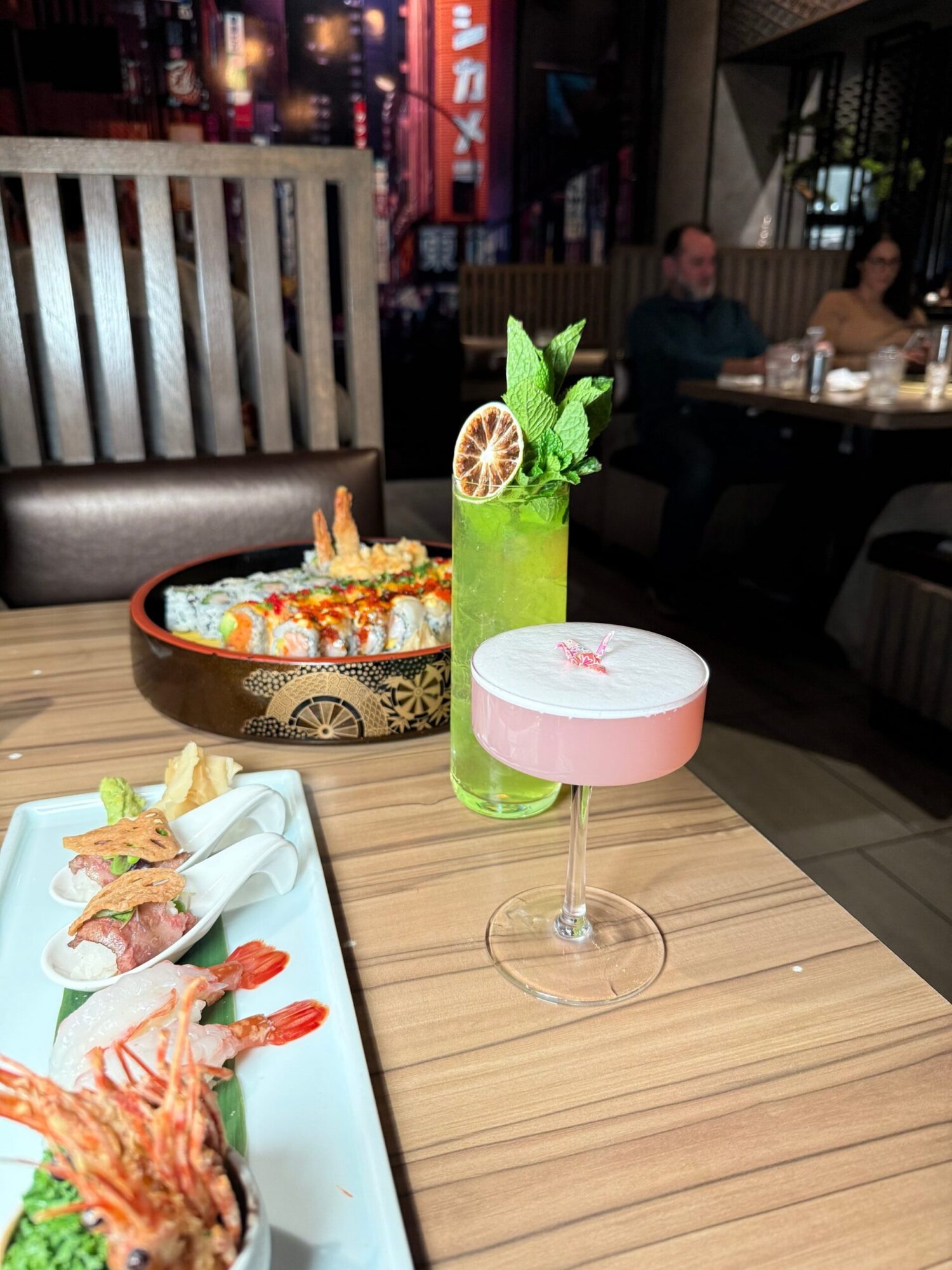 A green cocktail with garnishes, a sushi platter, and a pink drink on a wooden table in a restaurant.