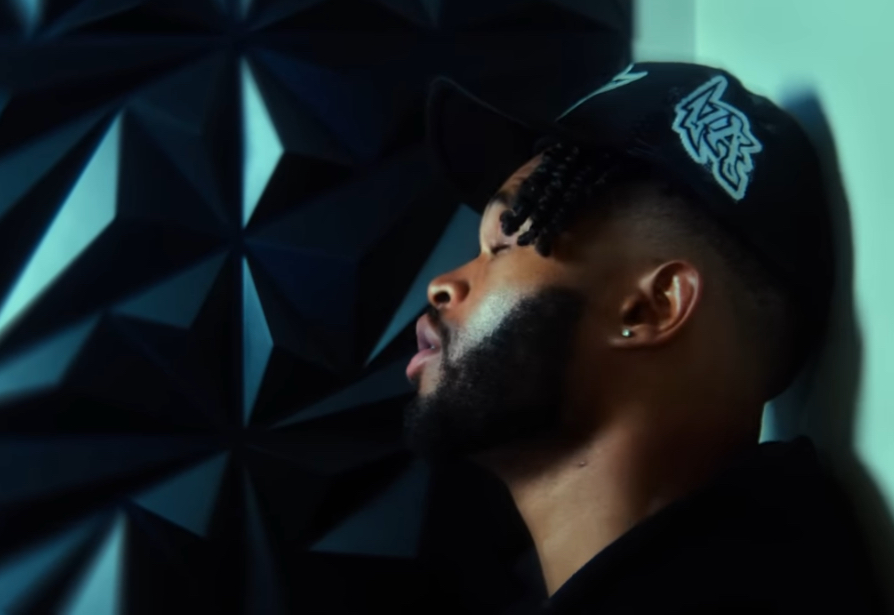 Side profile of a man with a beard wearing a black cap, leaning against a wall, with geometric patterns in the background.