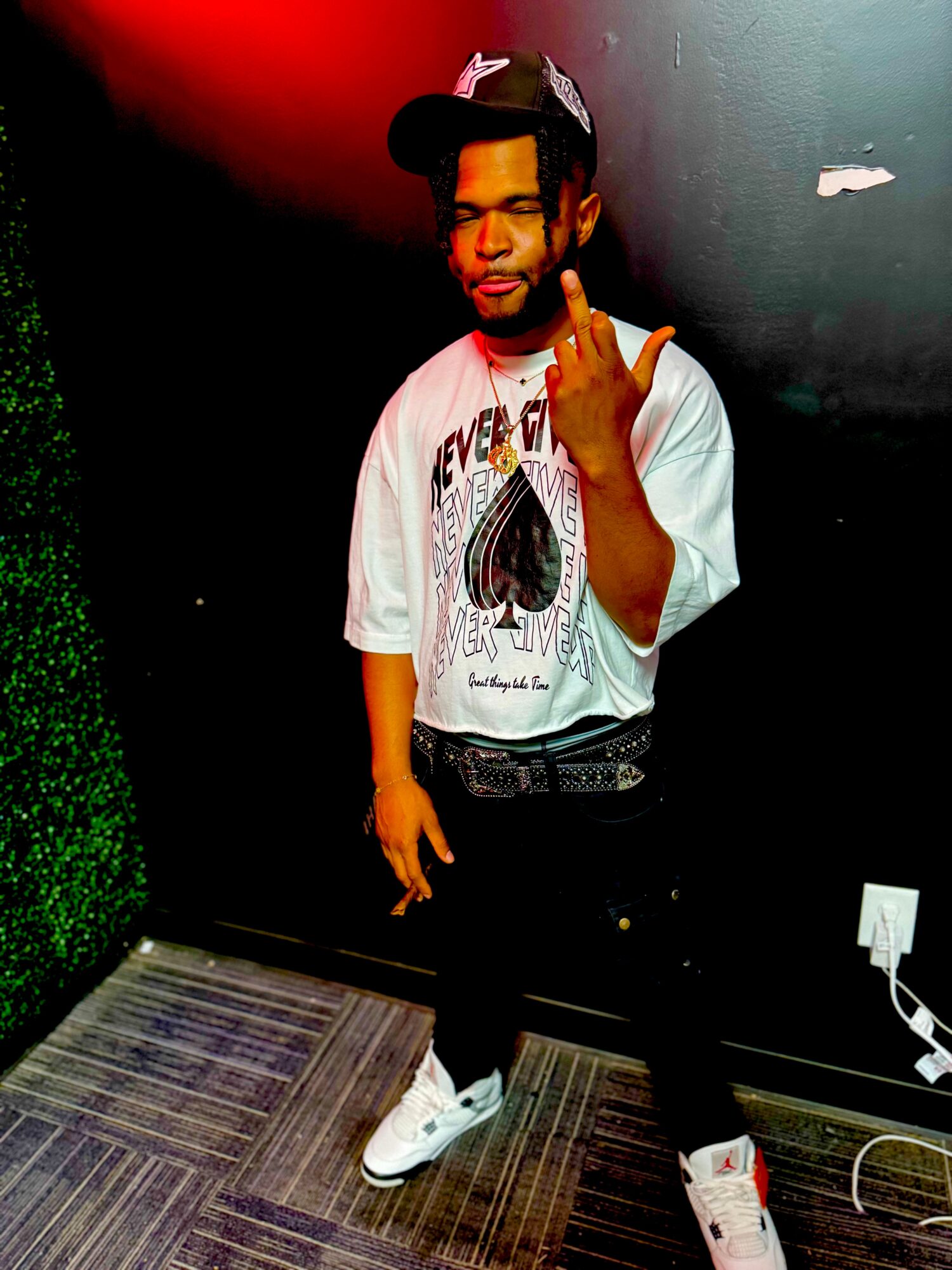 Young man with a cap, wearing a graphic t-shirt, making a hand gesture, standing indoors with a dark wall and green decor.
