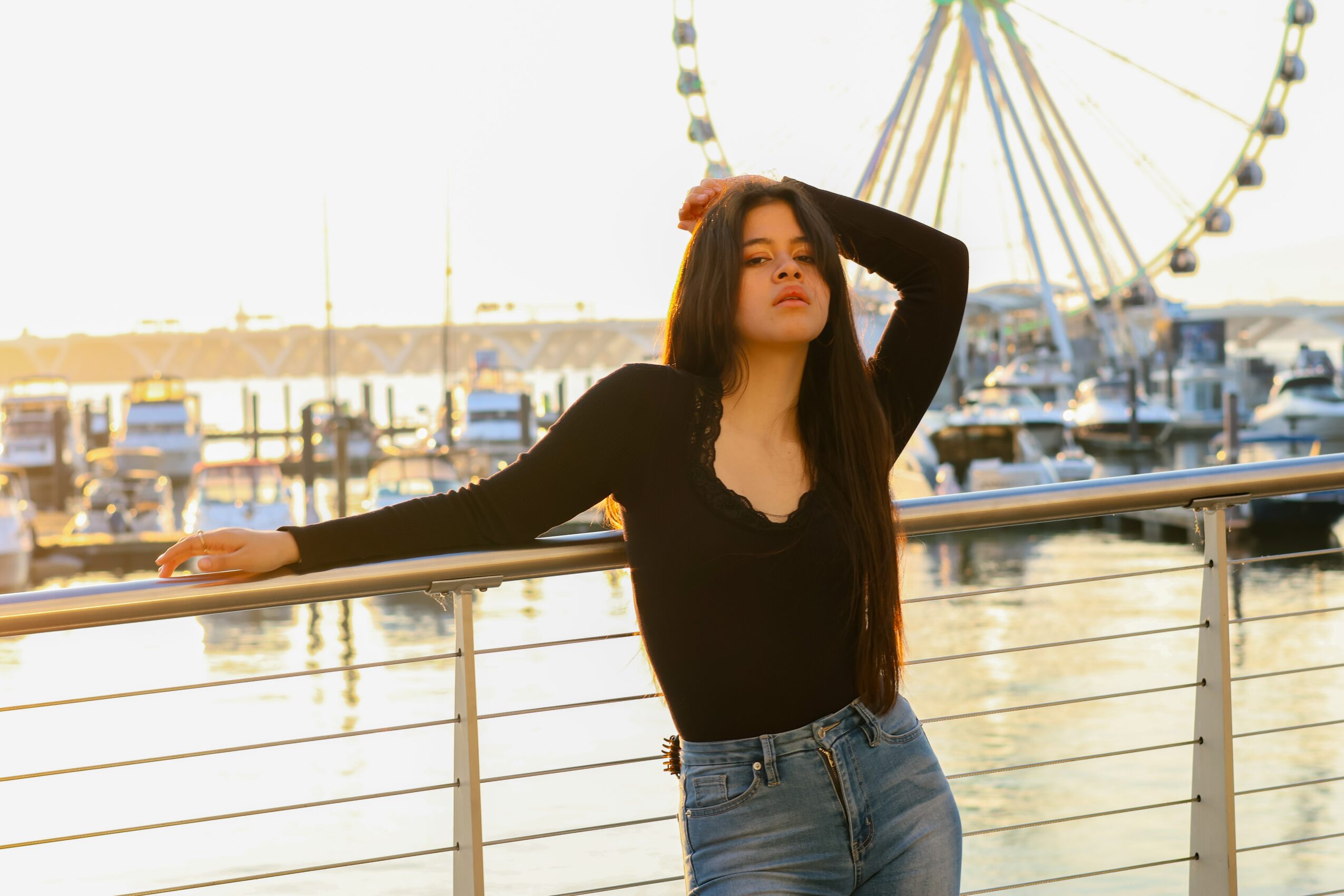 Young woman with long dark hair leaning on a railing at a marina, with a Ferris wheel in the background.