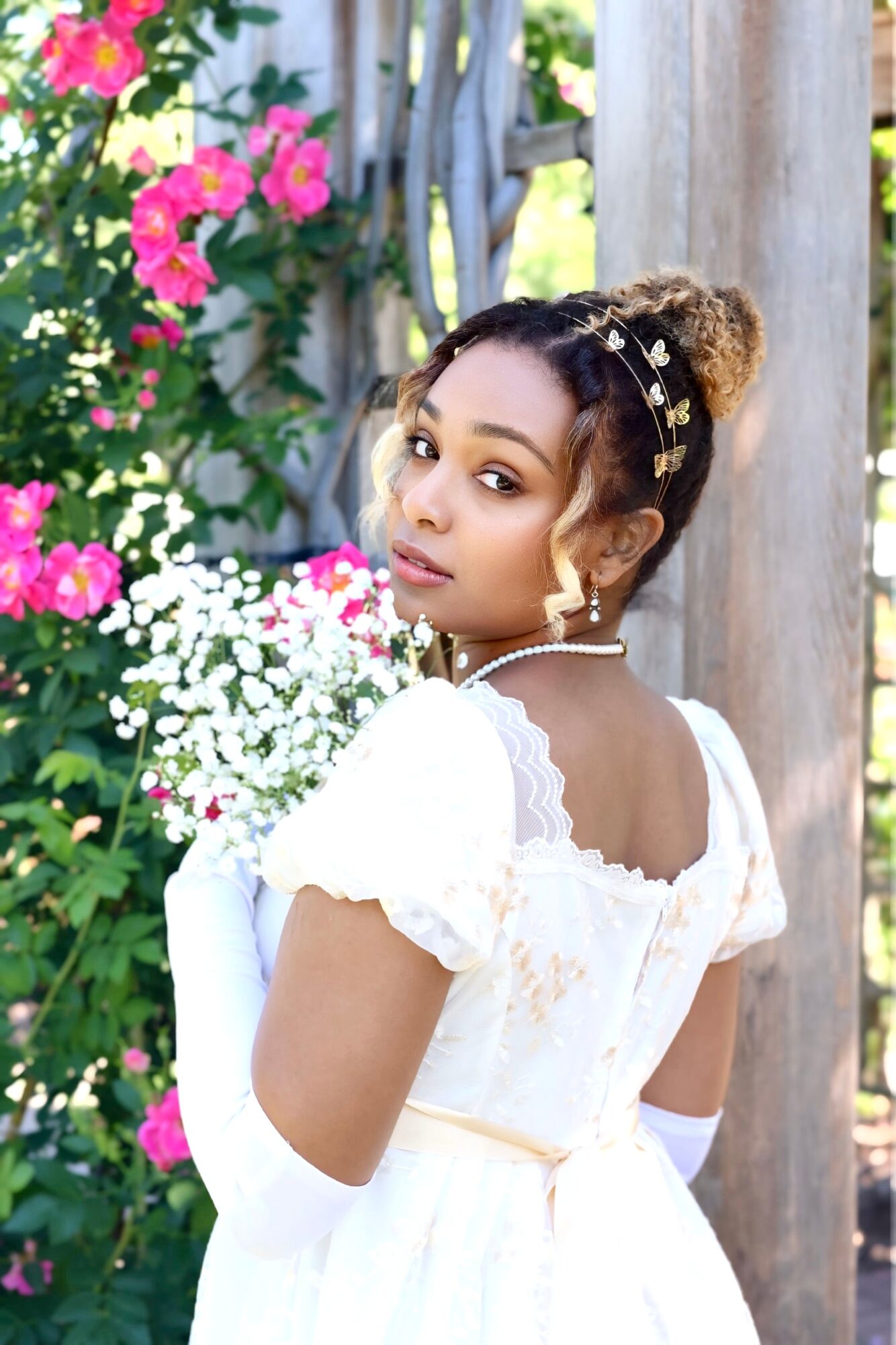 Young woman in white dress with floral background, looking over shoulder, with styled hair and accessories.