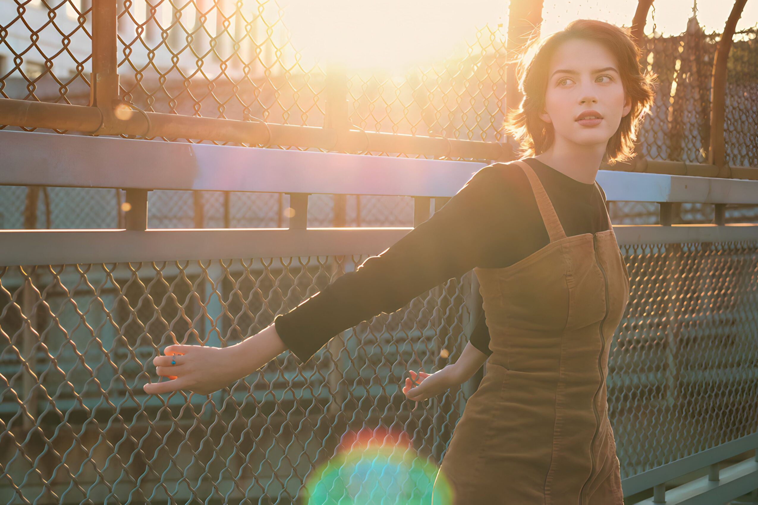 Young woman with short hair standing near a chain-link fence during sunset, looking to the side with outstretched arms.