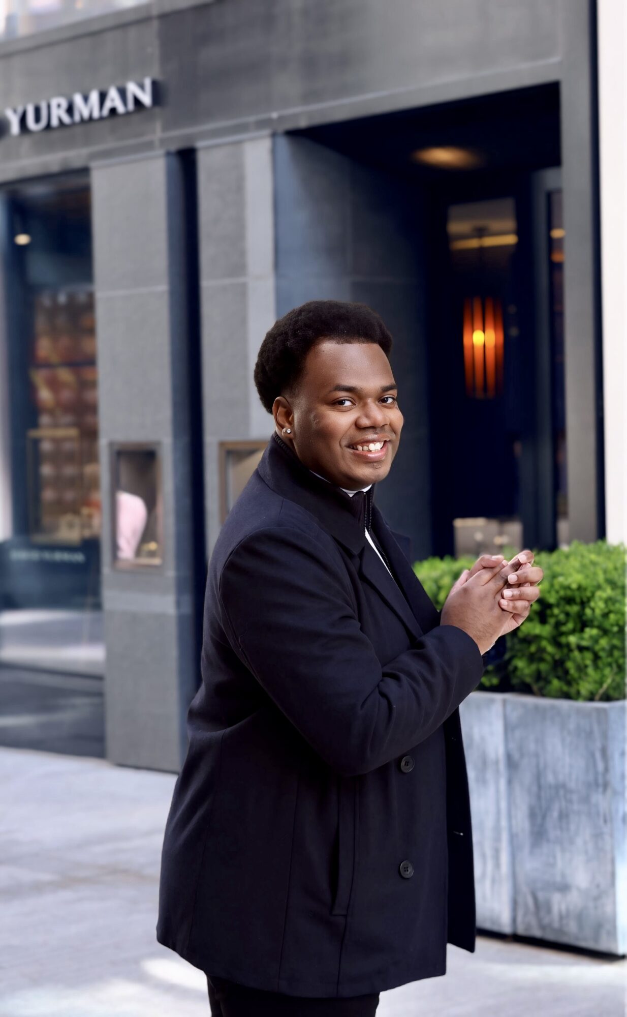 Young man smiling outdoors, standing near a building with a large planter, wearing a dark jacket, holding a phone.