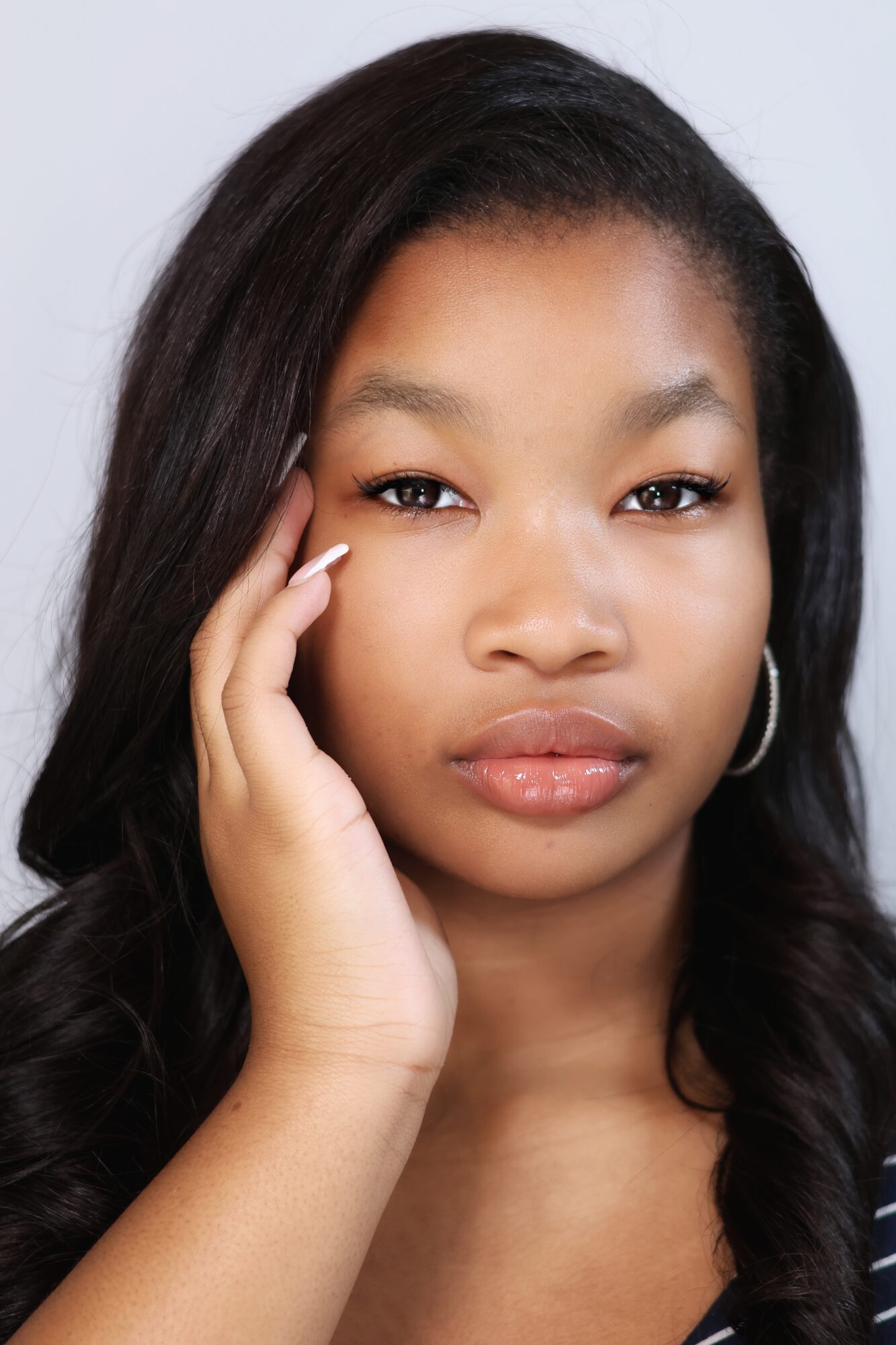 Young woman with long dark hair, touching her face gently, looking directly at the camera, neutral expression.