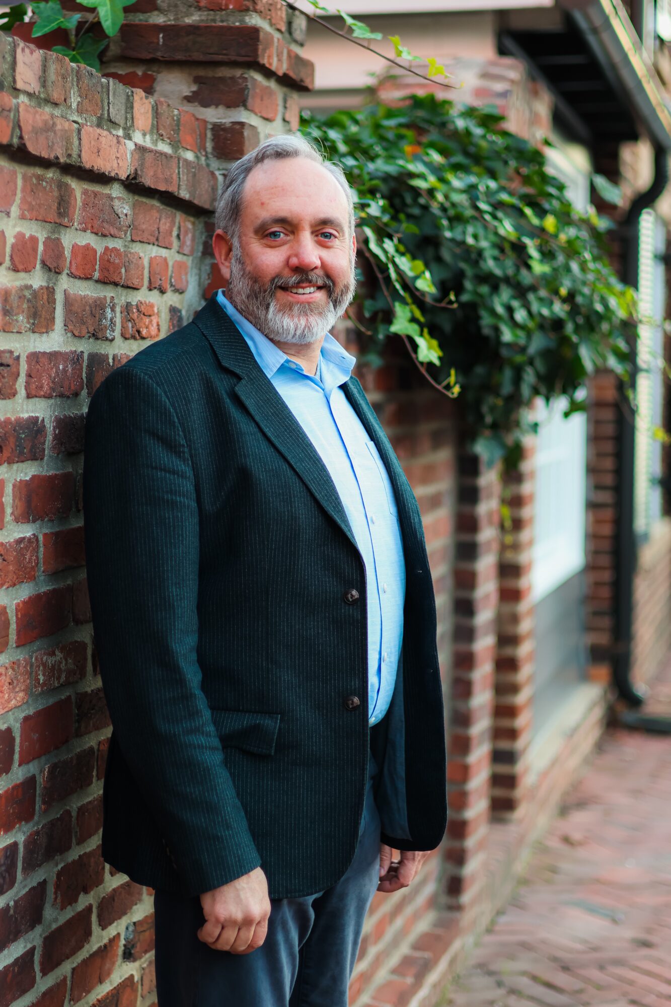 Man with gray hair and beard smiling, standing against a brick wall with greenery in the background.