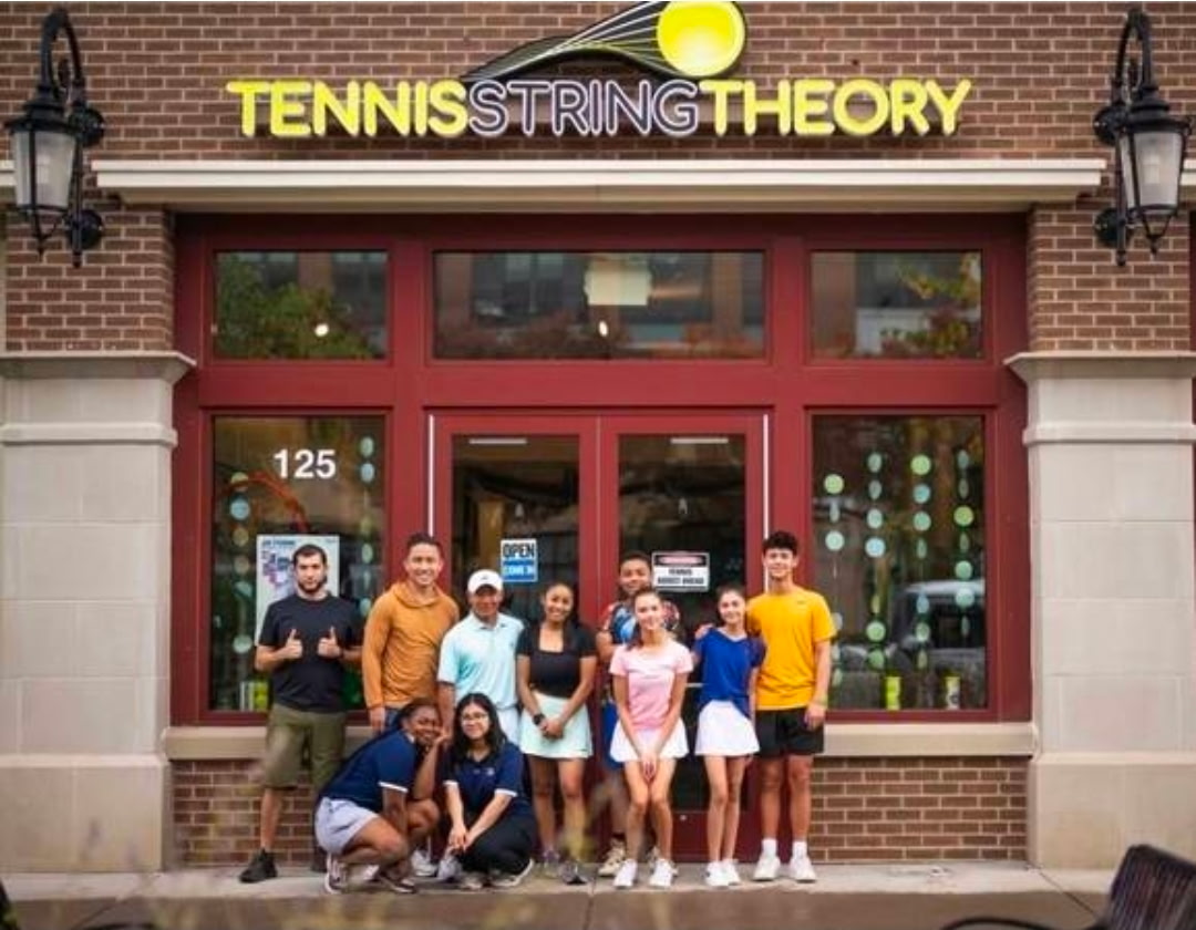 Group of nine people standing in front of a tennis shop with a sign reading 'Tennis String Theory'.