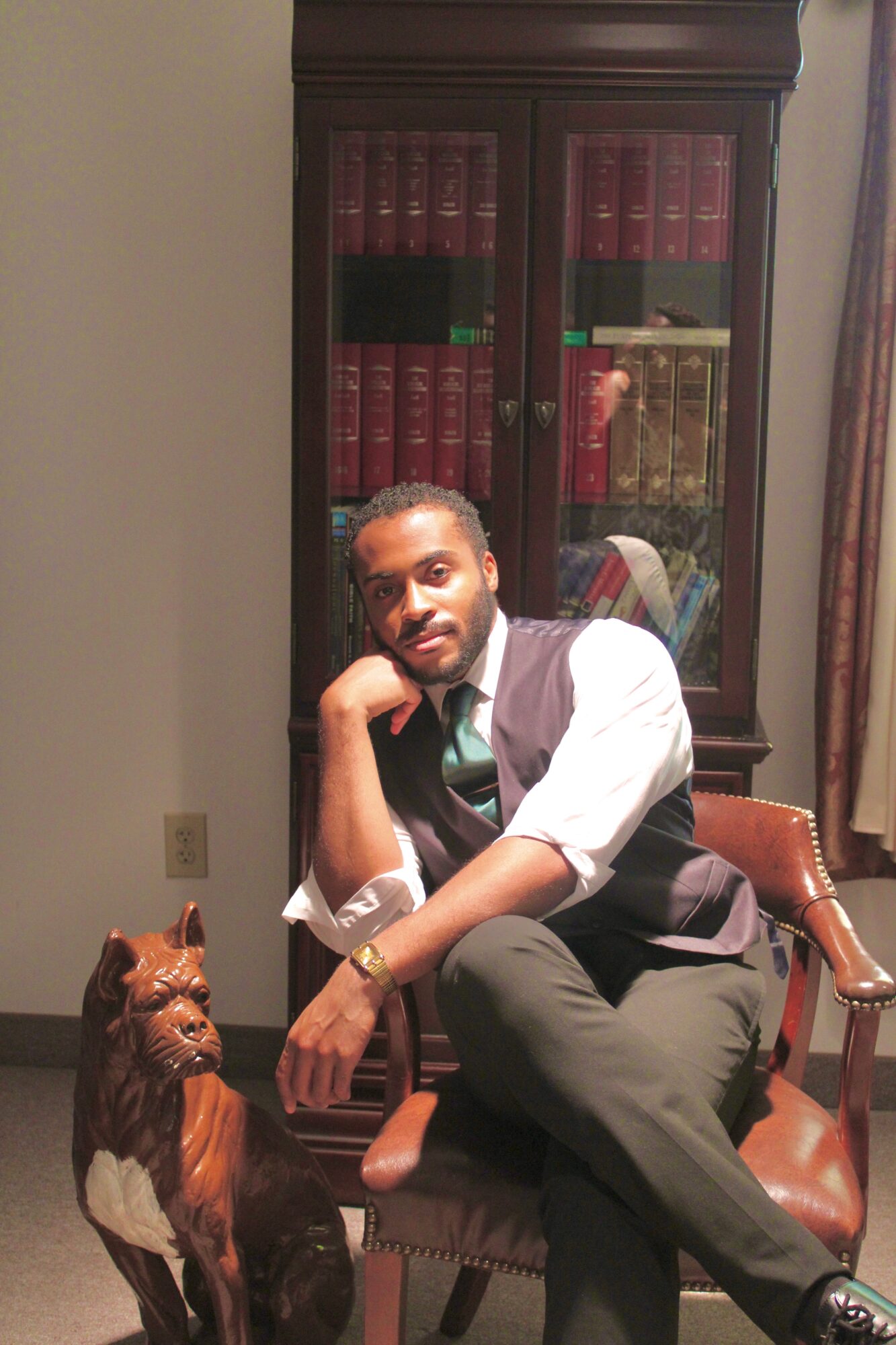 Man sitting on a wooden chair with a leather seat, in a room with a bookshelf and curtains, resting his chin on his hand.