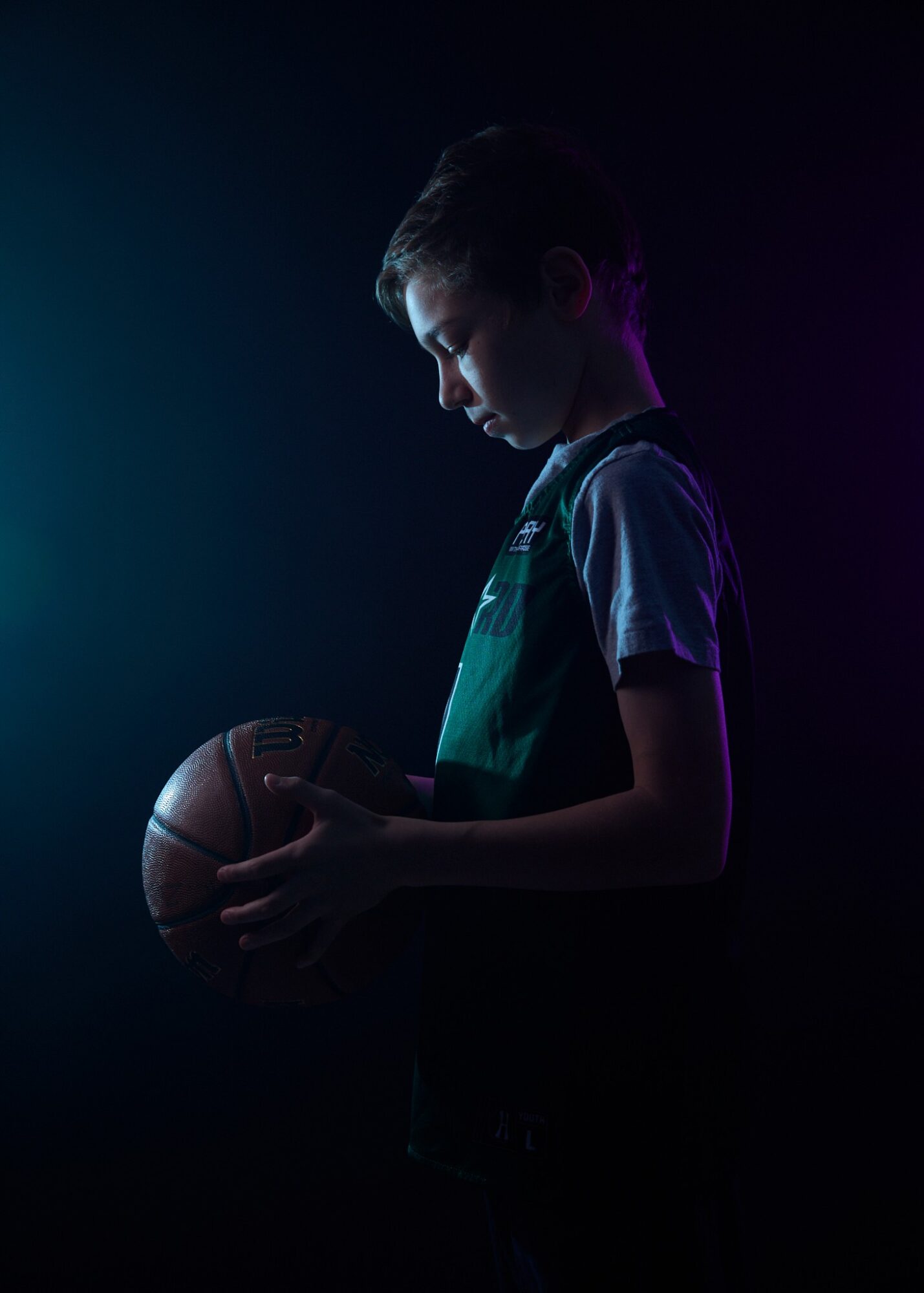 Child holding a basketball in a dark setting with subtle lighting on the side, looking down at the ball.