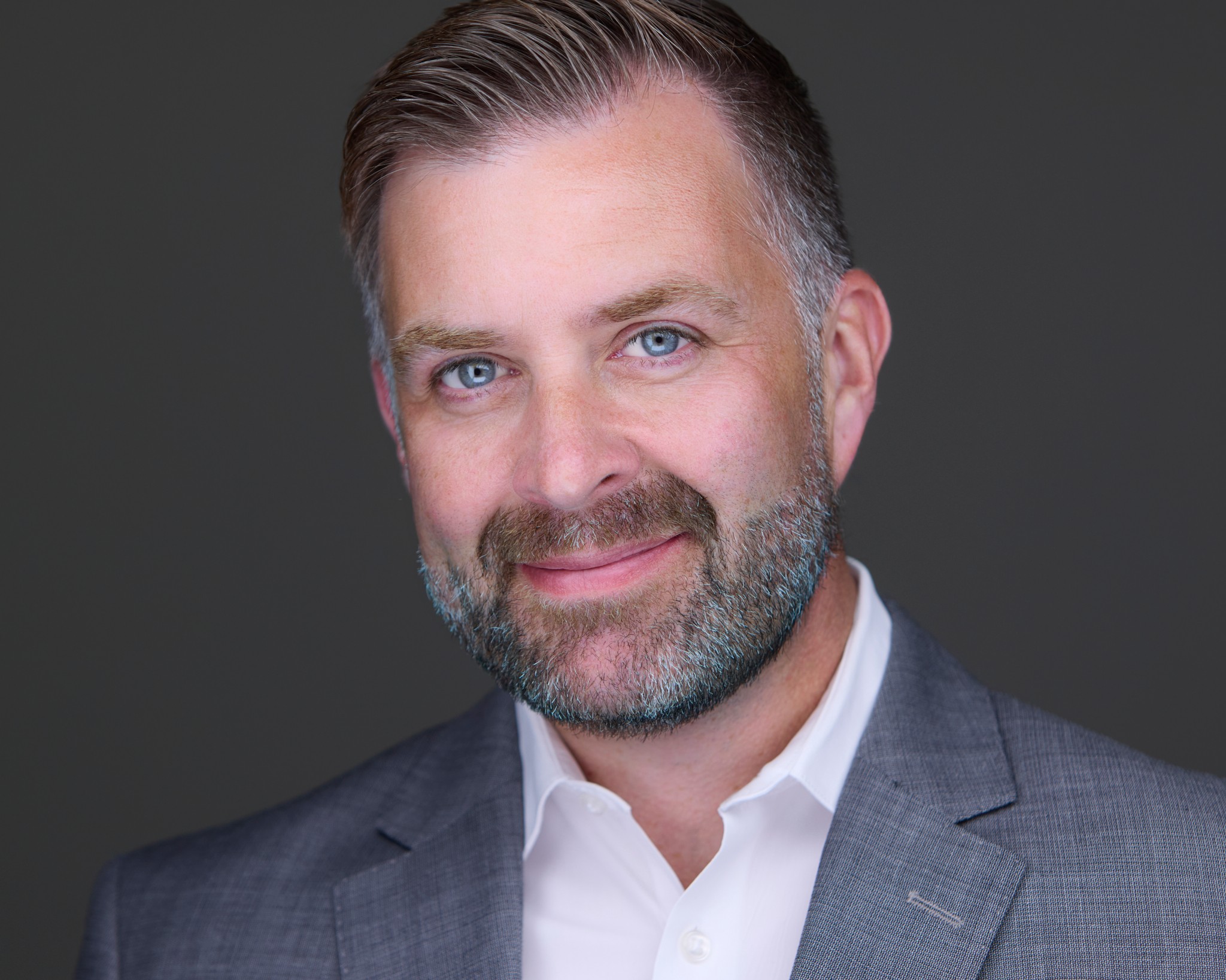 Portrait of a man with short hair, beard, wearing a suit and white shirt, smiling against a dark background.