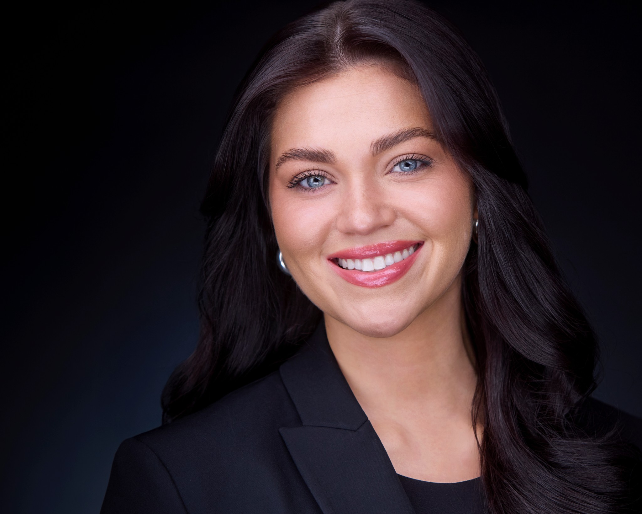 Young woman with long dark hair, smiling, wearing a black blazer, against a dark background.