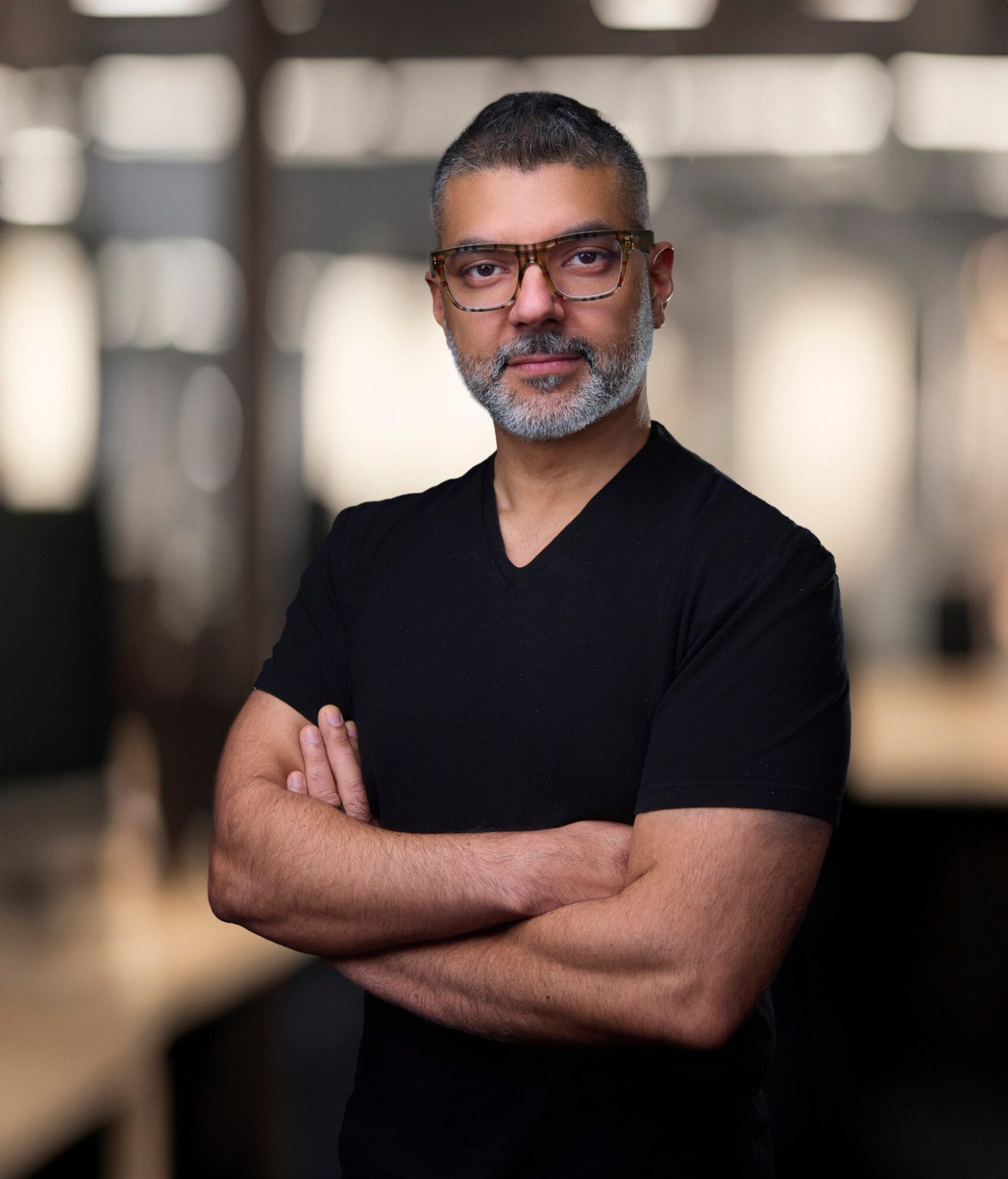 Man with glasses and beard wearing a black shirt, arms crossed, in an indoor setting with blurred background.
