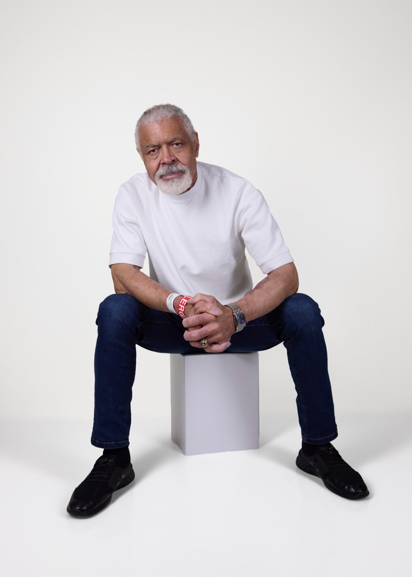 Older man with gray hair and beard sitting on a white cube, smiling, wearing a white t-shirt and dark jeans.