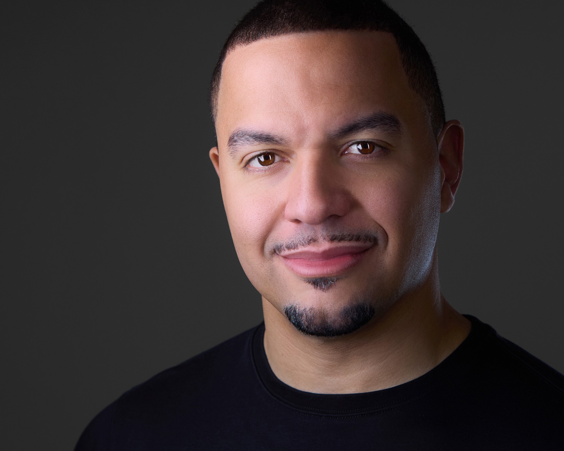 Portrait of a man with short dark hair, slight beard, smiling, wearing a black shirt, dark background.