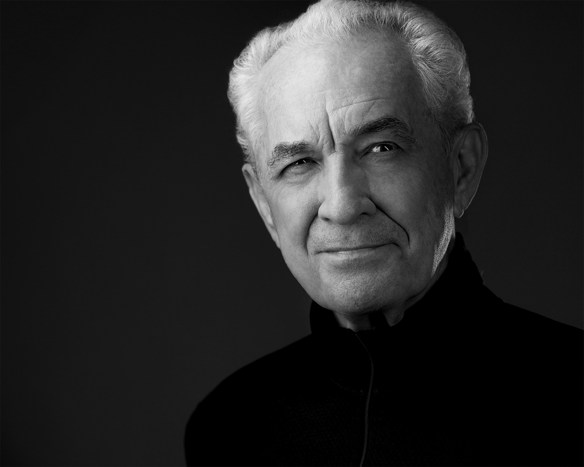 Portrait of an elderly man with white hair, wearing a dark turtleneck, looking at the camera with a slight smile.