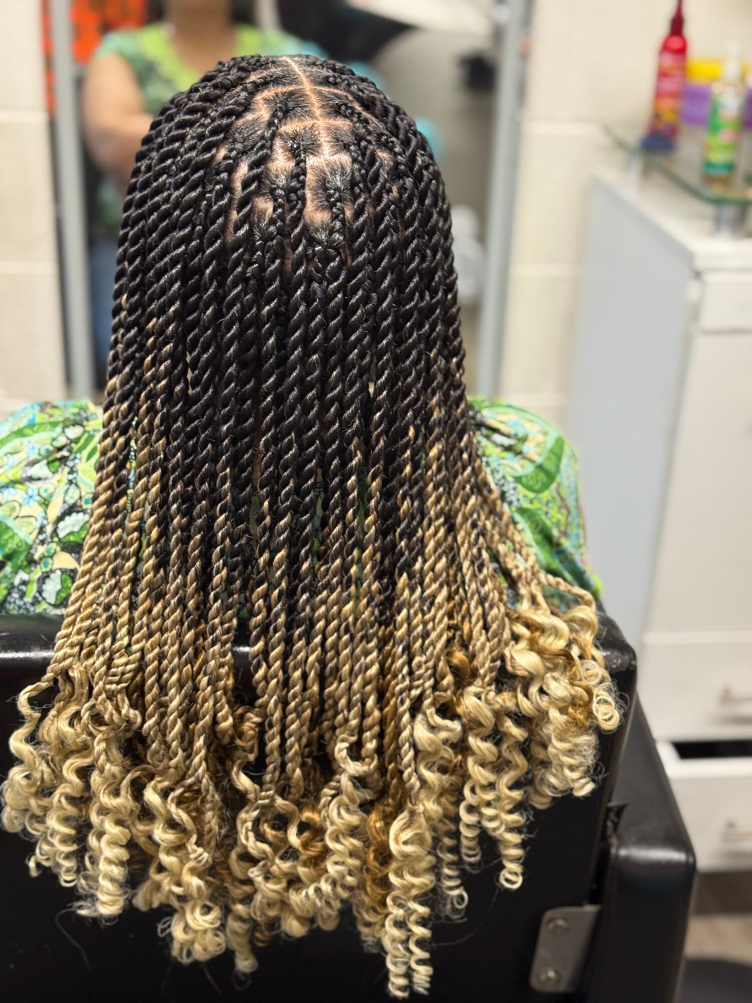 Back view of a person with long, twisted braids transitioning from dark roots to blonde ends, wearing a green patterned top.
