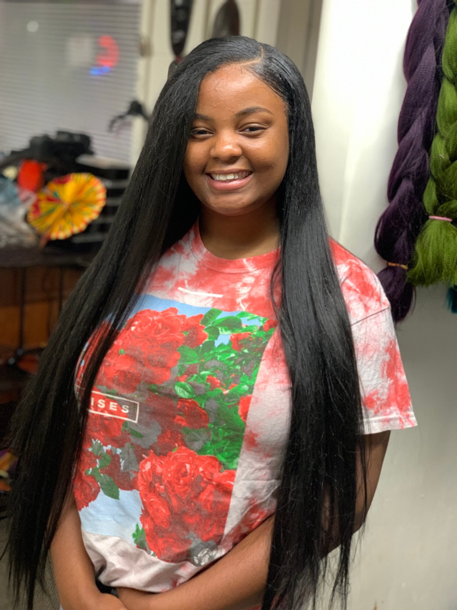 Smiling young woman with long, straight black hair wearing a colorful floral T-shirt, standing indoors.