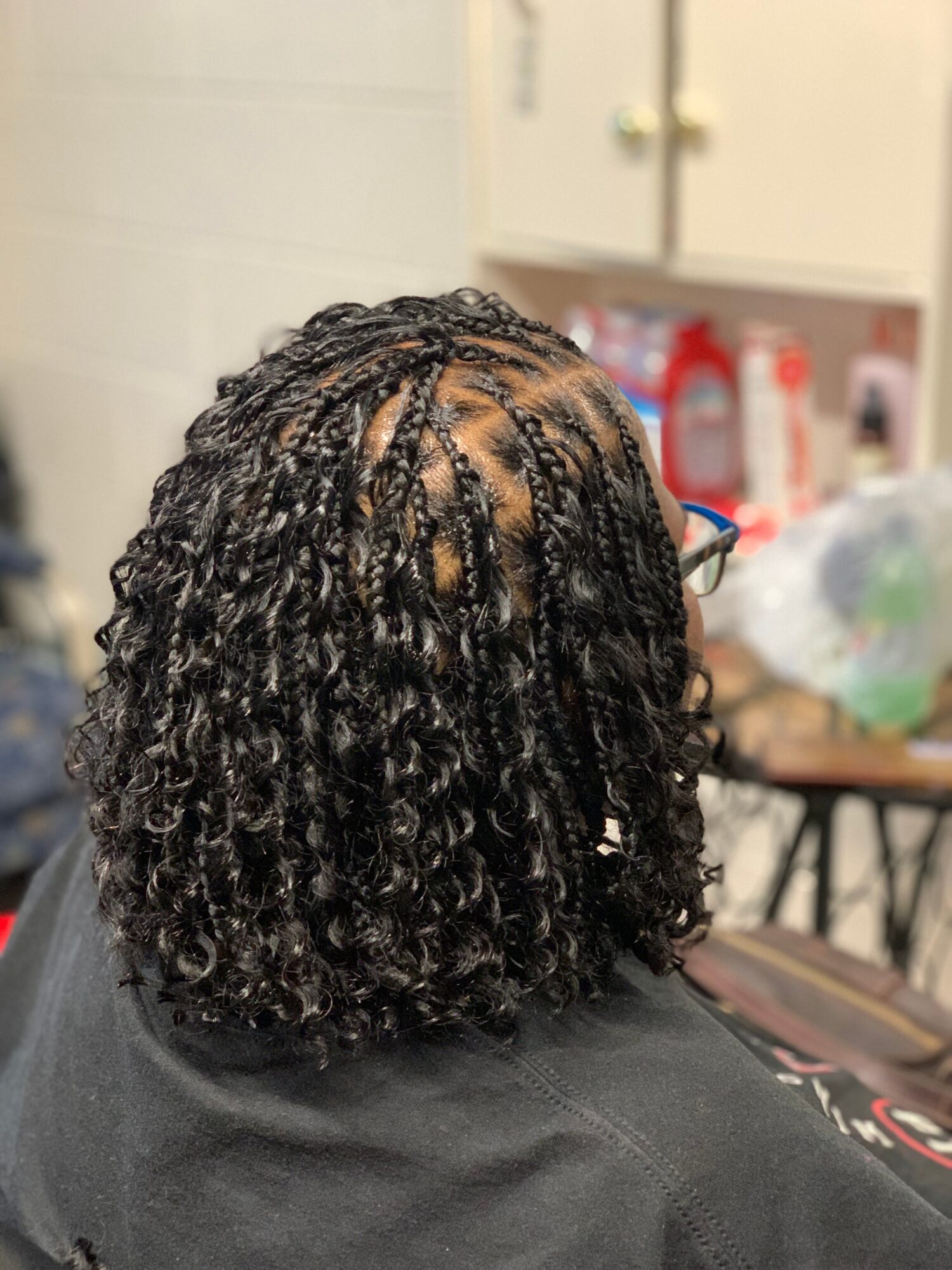 Back view of person with short, curly, braided hair, wearing glasses, in an indoor setting with shelves and objects in background.