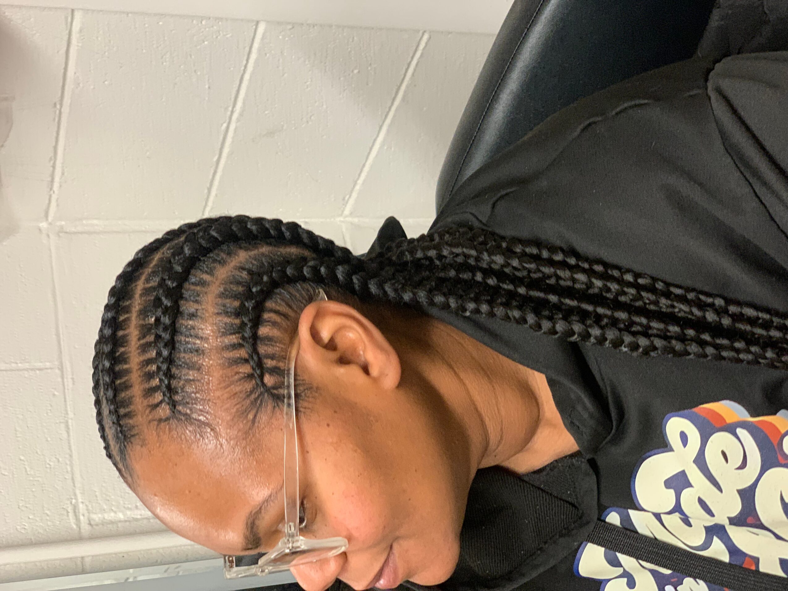 Person with braided hair and glasses, wearing a black shirt, sitting indoors against a tiled wall.