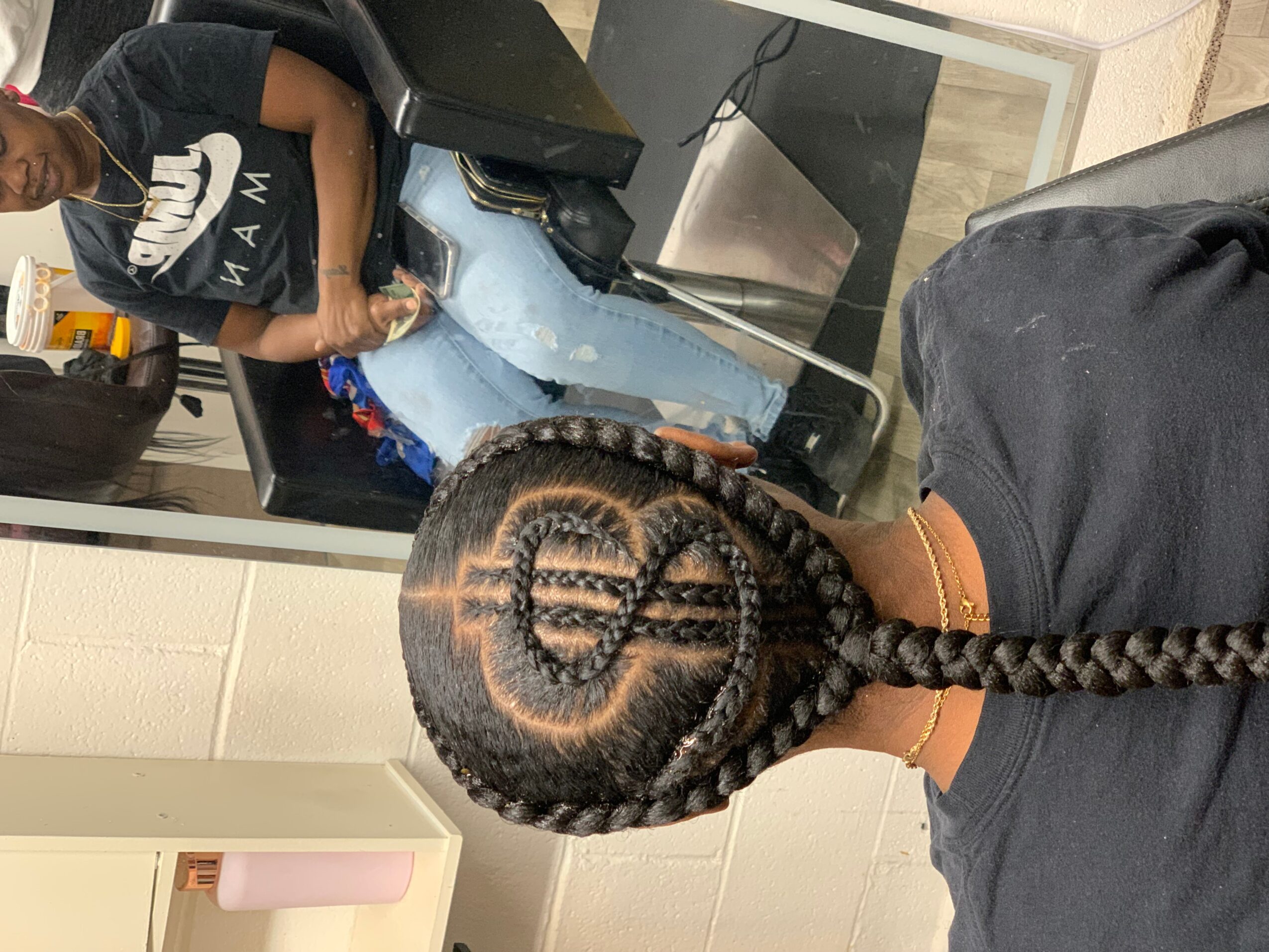 Back of a person with braided hair featuring a design of a dollar sign, in a room with a person sitting at a desk.