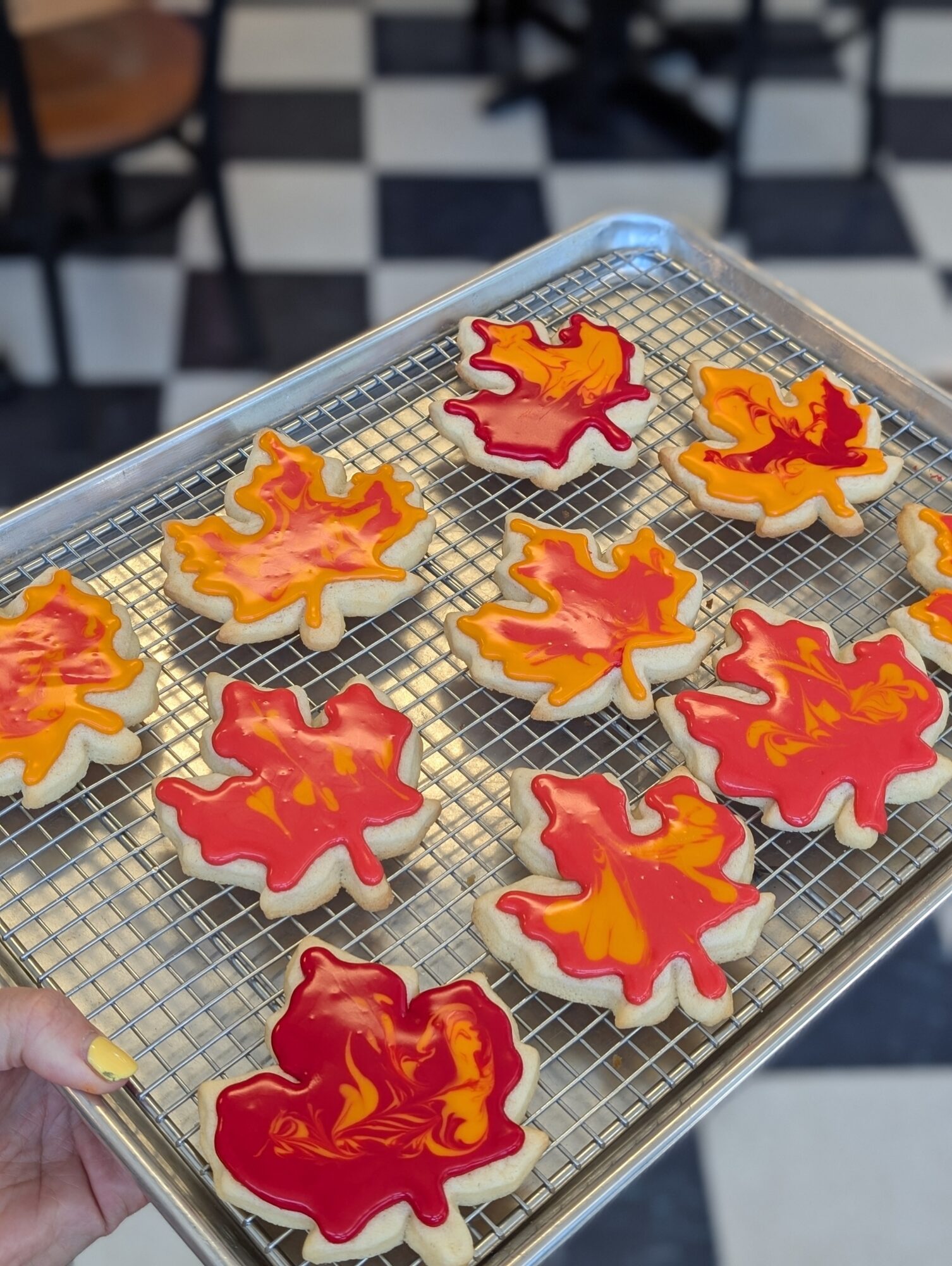 Tray of decorated cookies with red and orange leaf shapes on a wire rack, background of black and white checkered floor.
