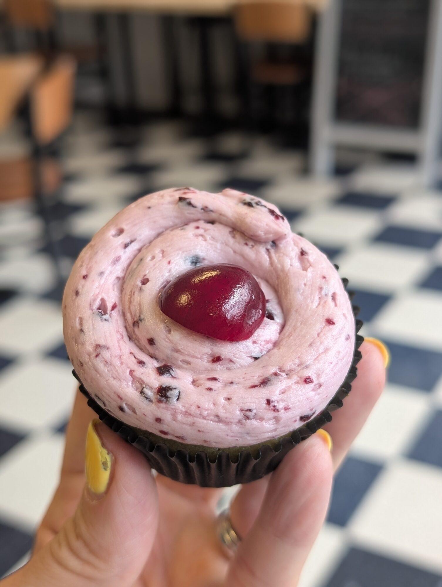 Cupcake with pink frosting and a cherry on top, held by a person with yellow nail polish, in a checkered floor setting.