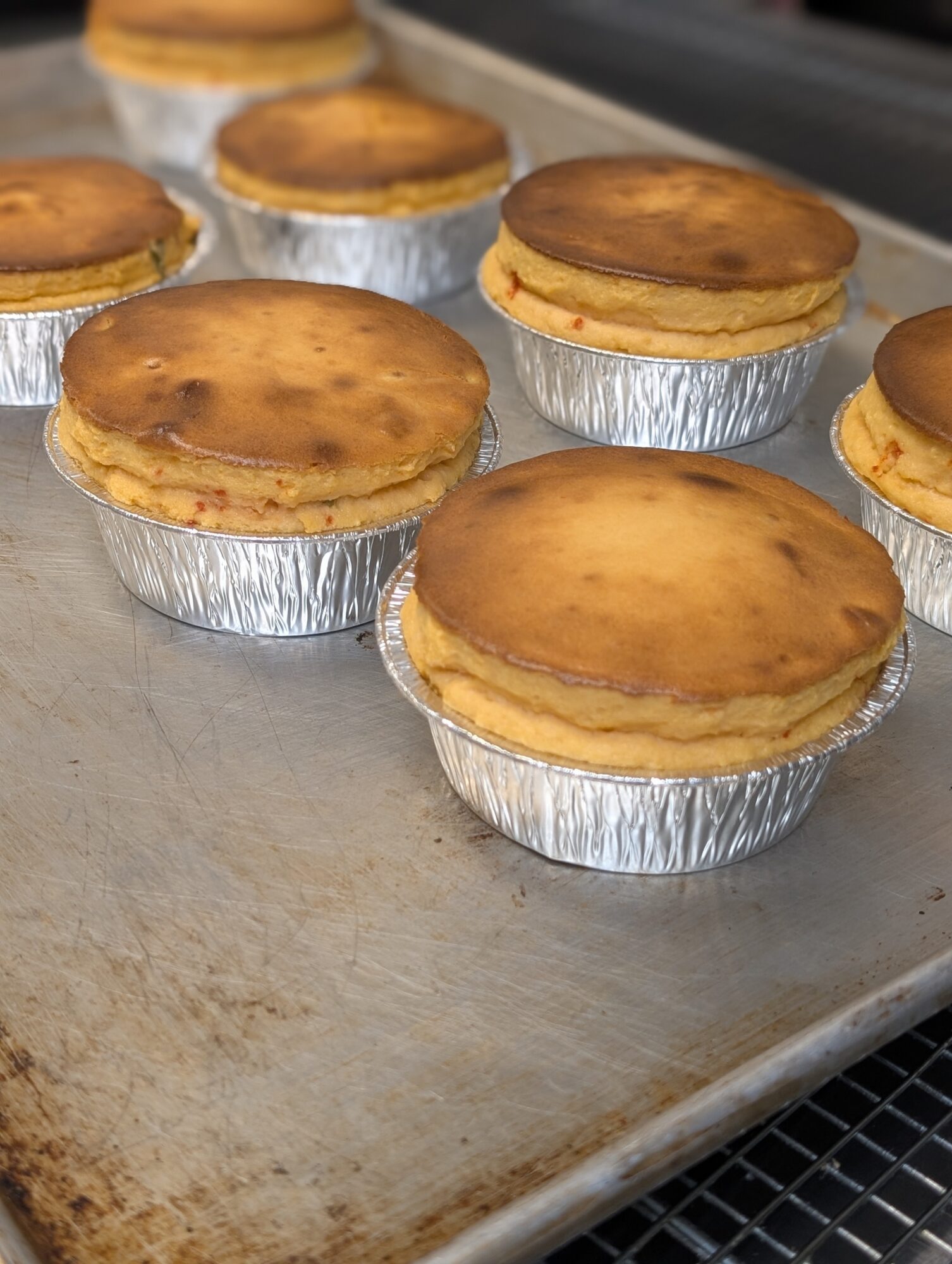 Several baked cheesecakes in foil cups on a metal baking sheet, with a golden-brown top.