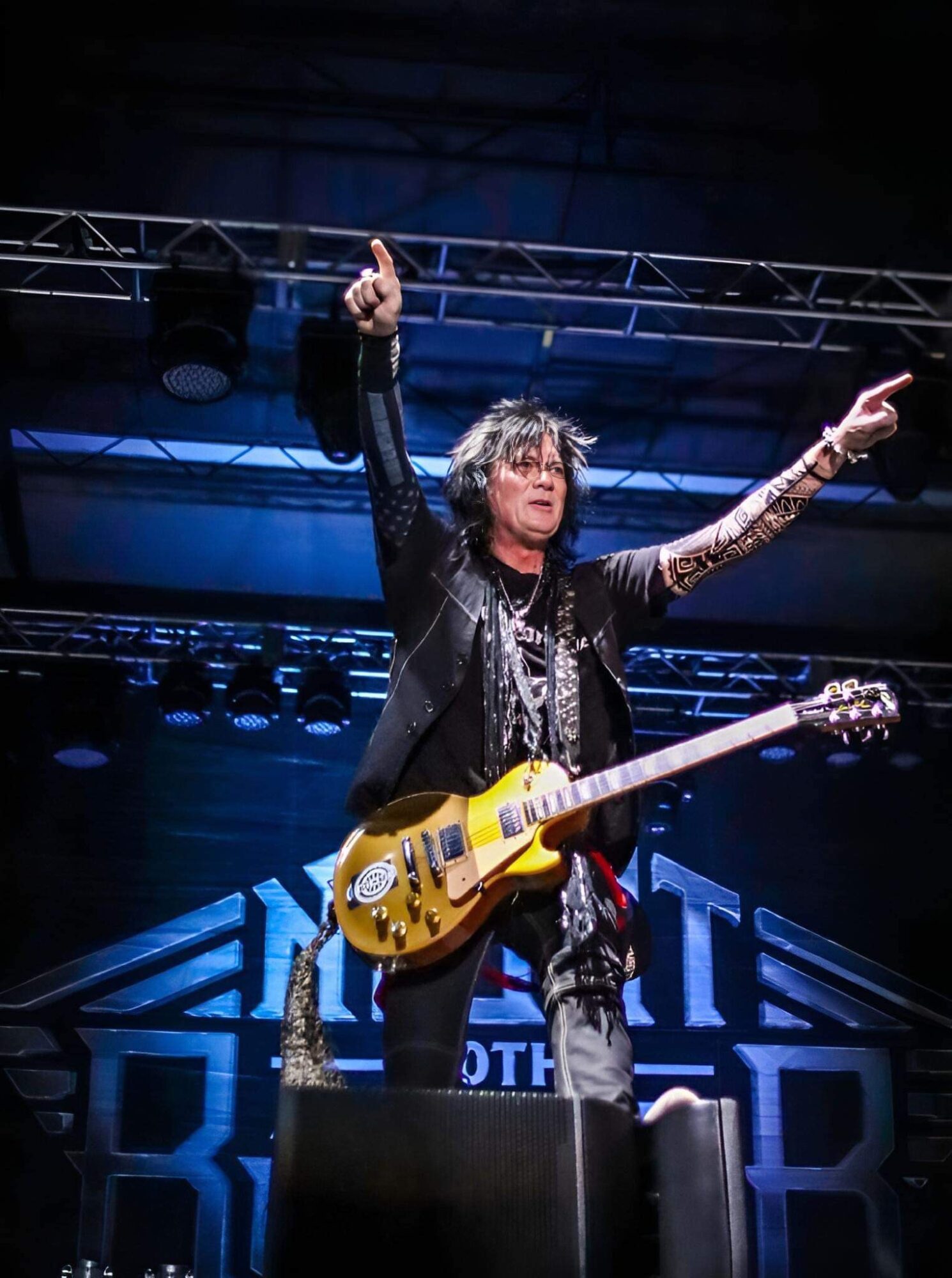 Musician on stage holding a yellow electric guitar, pointing upward with one hand, with stage lights and backdrop behind.