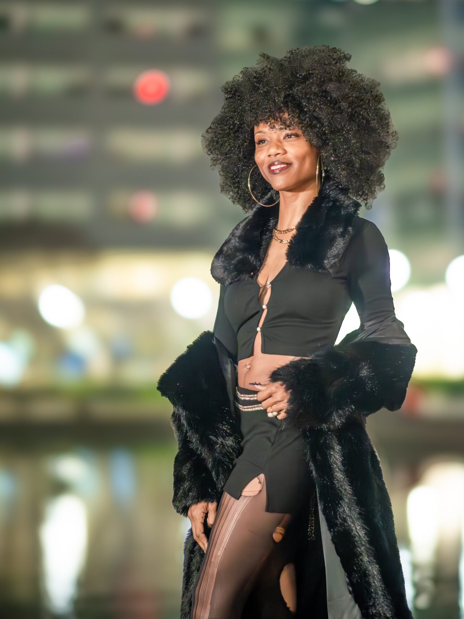 Woman with curly hair in a black outfit and fur coat, standing outdoors with blurred city lights background.