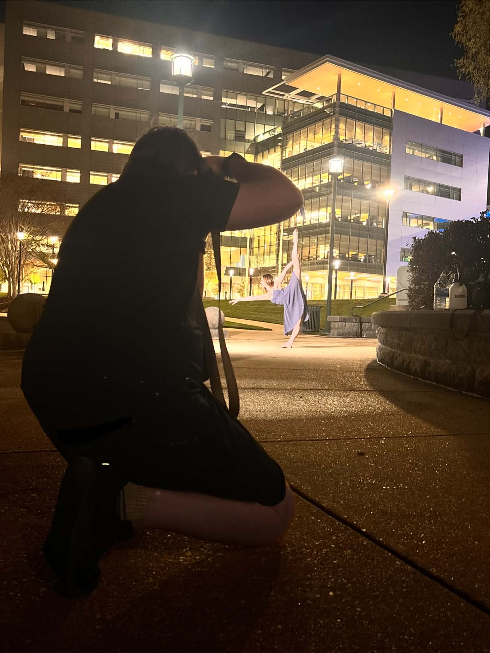 Person kneeling on the ground taking a photo of a building at night, illuminated with bright lights.