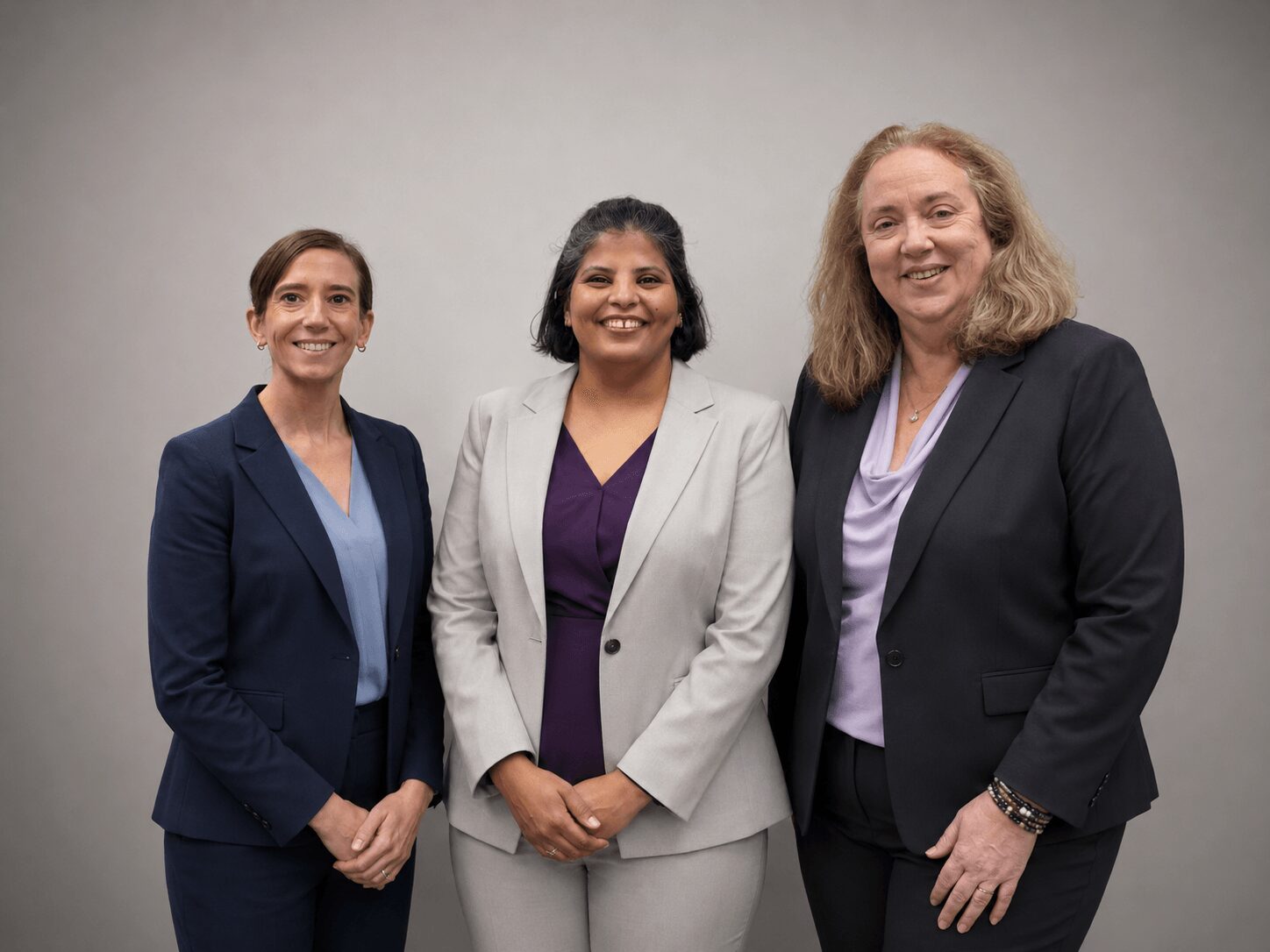 Three women in business attire standing together, smiling, against a plain background.