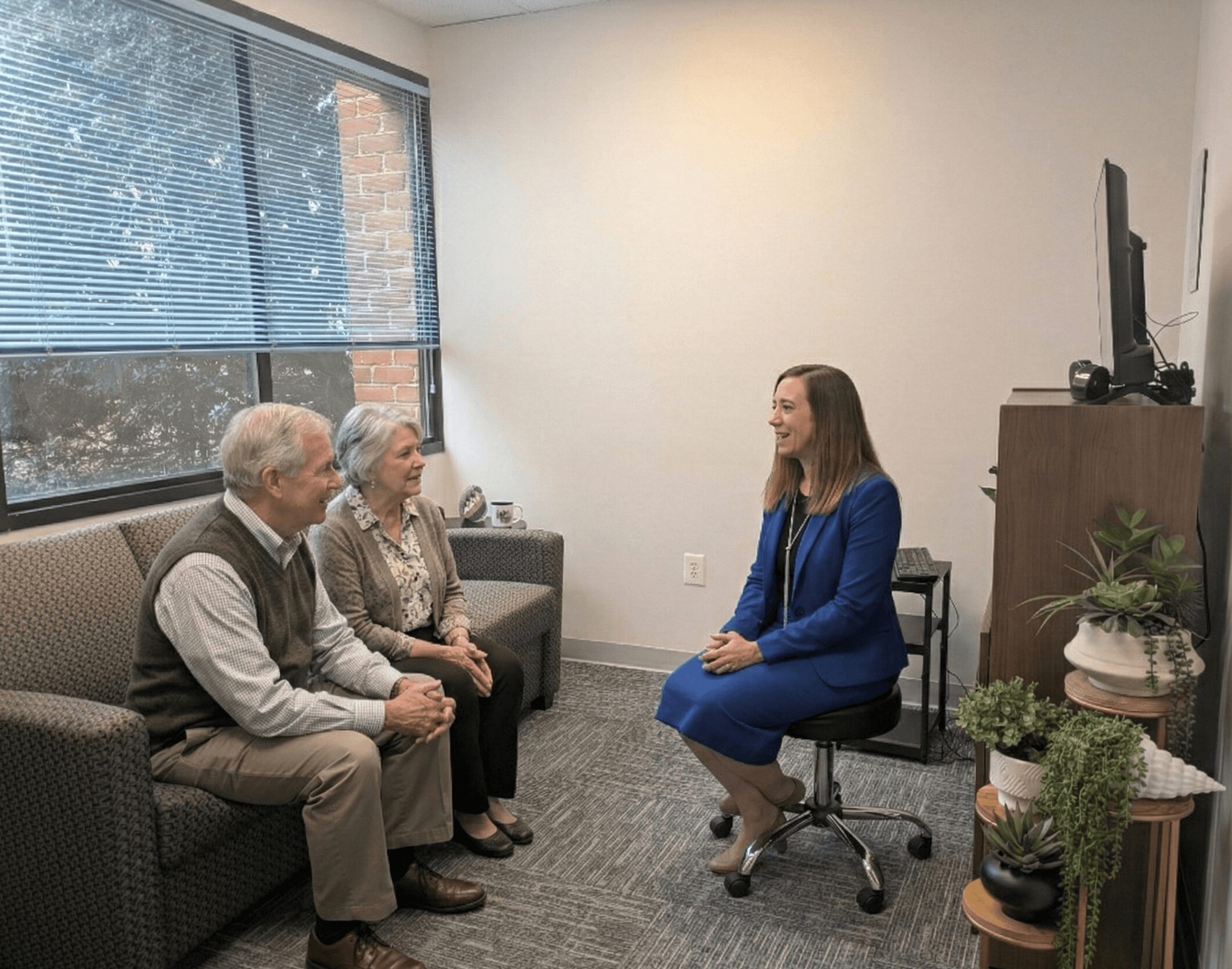 Three older adults sitting on a sofa and a woman in a blue dress sitting on a chair in an office, near a window.