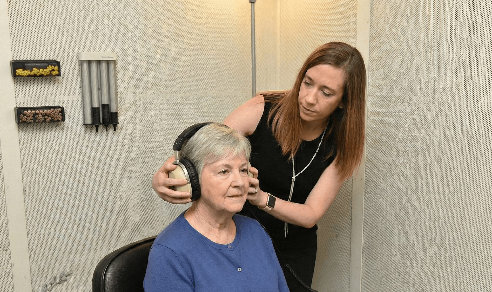 Young woman adjusting headphones on an elderly woman in a room with textured walls.