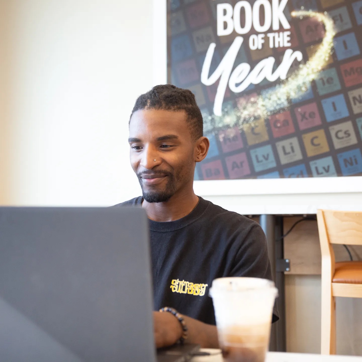 Man with braided hair working on a laptop in a cafe, with a poster reading 'Book of the Year' behind him.