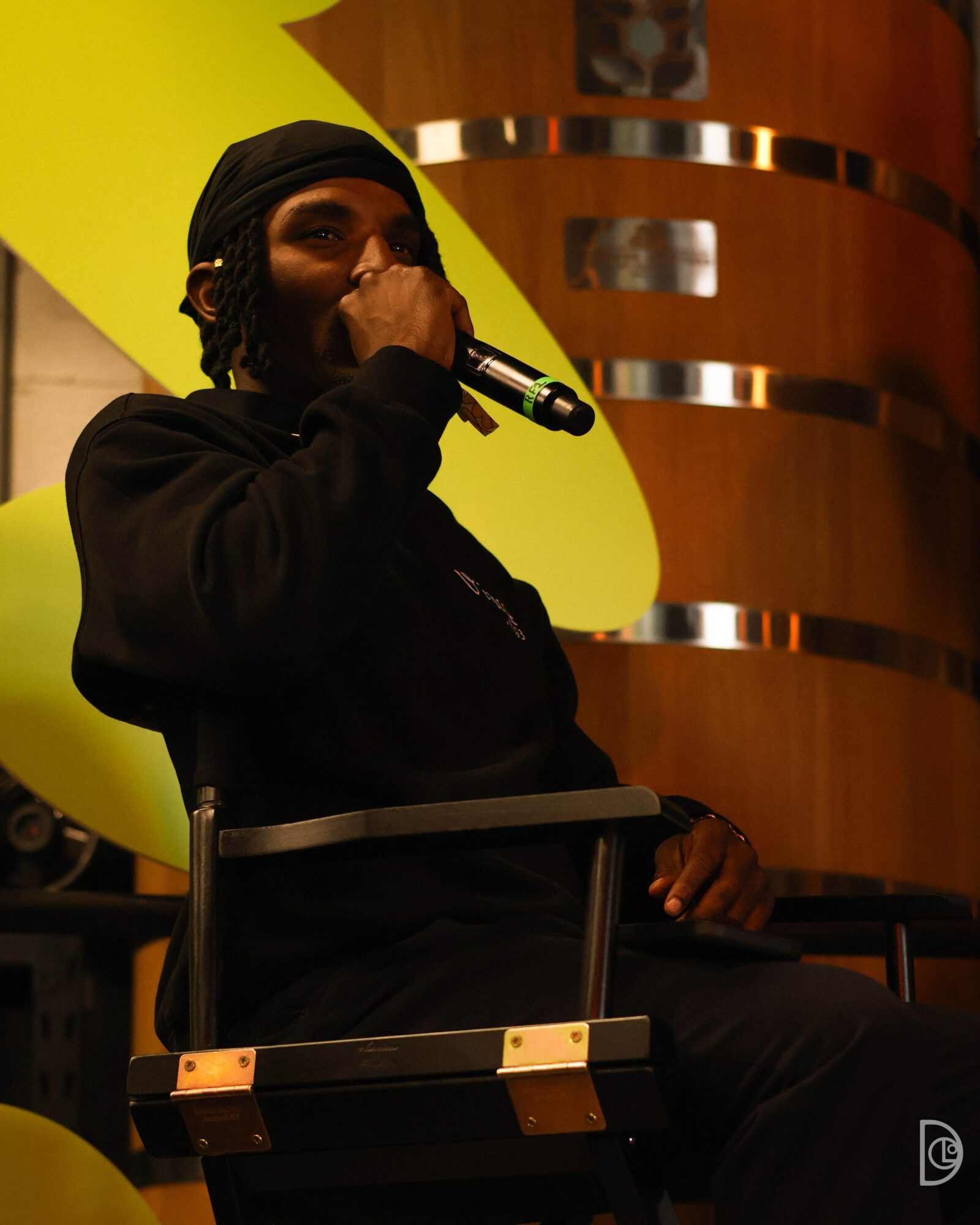Person wearing a black head covering and jacket, holding a microphone, sitting indoors with wooden and yellow background elements.