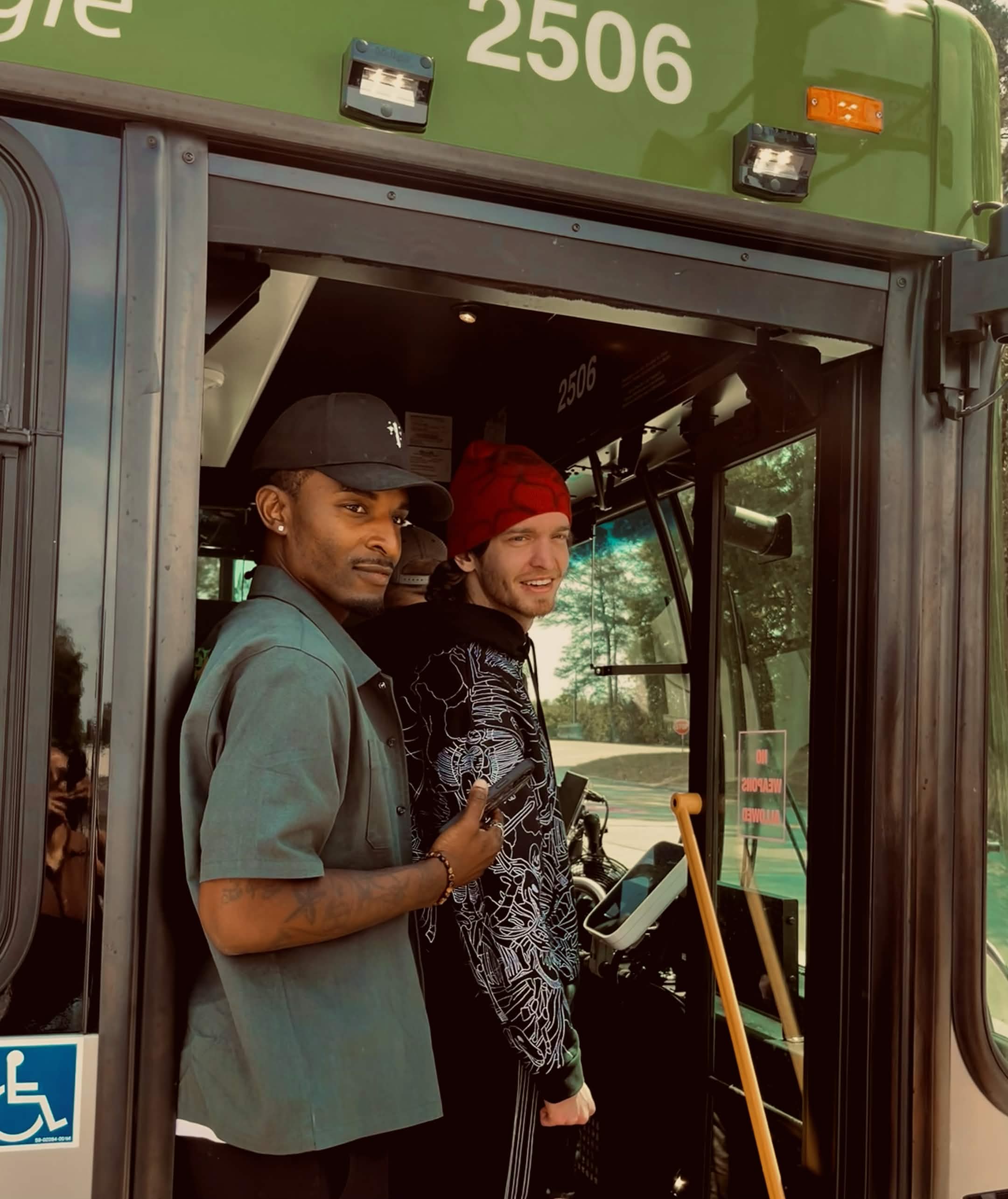 Two people standing inside a bus, smiling, with trees visible through the windows, one wearing a hat and the other a red beanie.