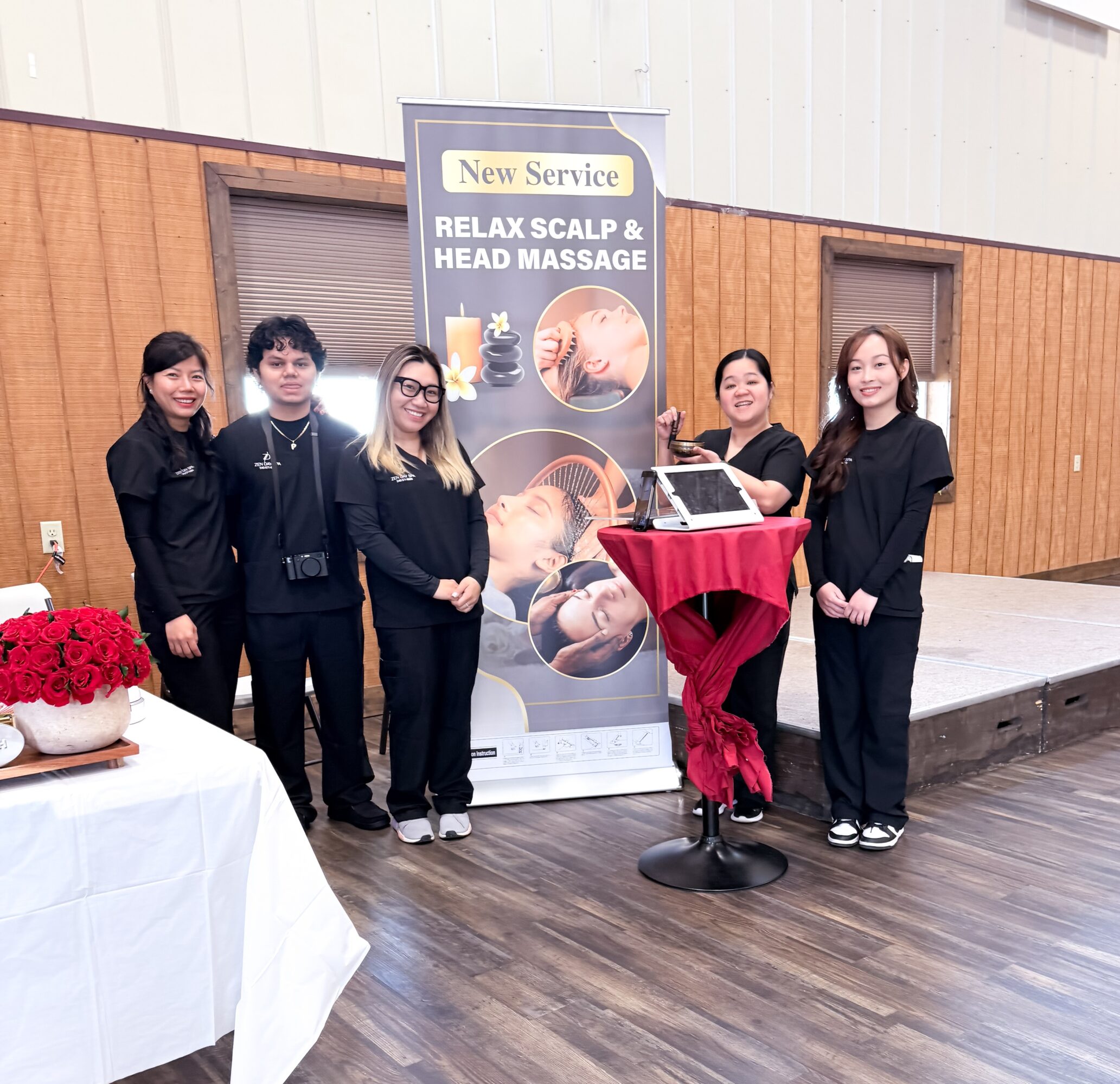 Five people stand next to a promotional banner for scalp and head massage services indoors, with a table of red flowers nearby.