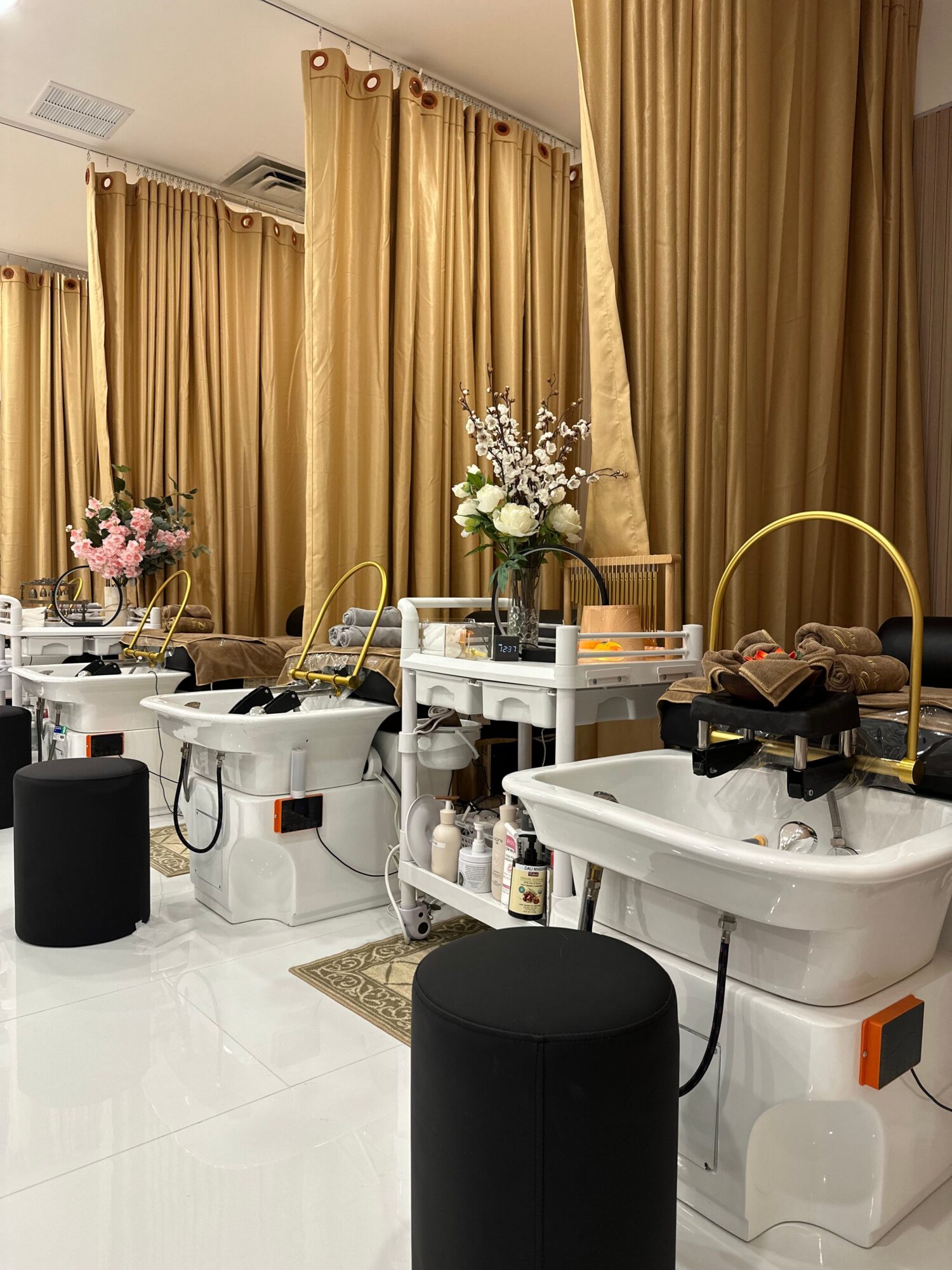 Table with flowers, bottles, and decorative items in front of gold curtains, with black and white furniture and a white tiled floor.