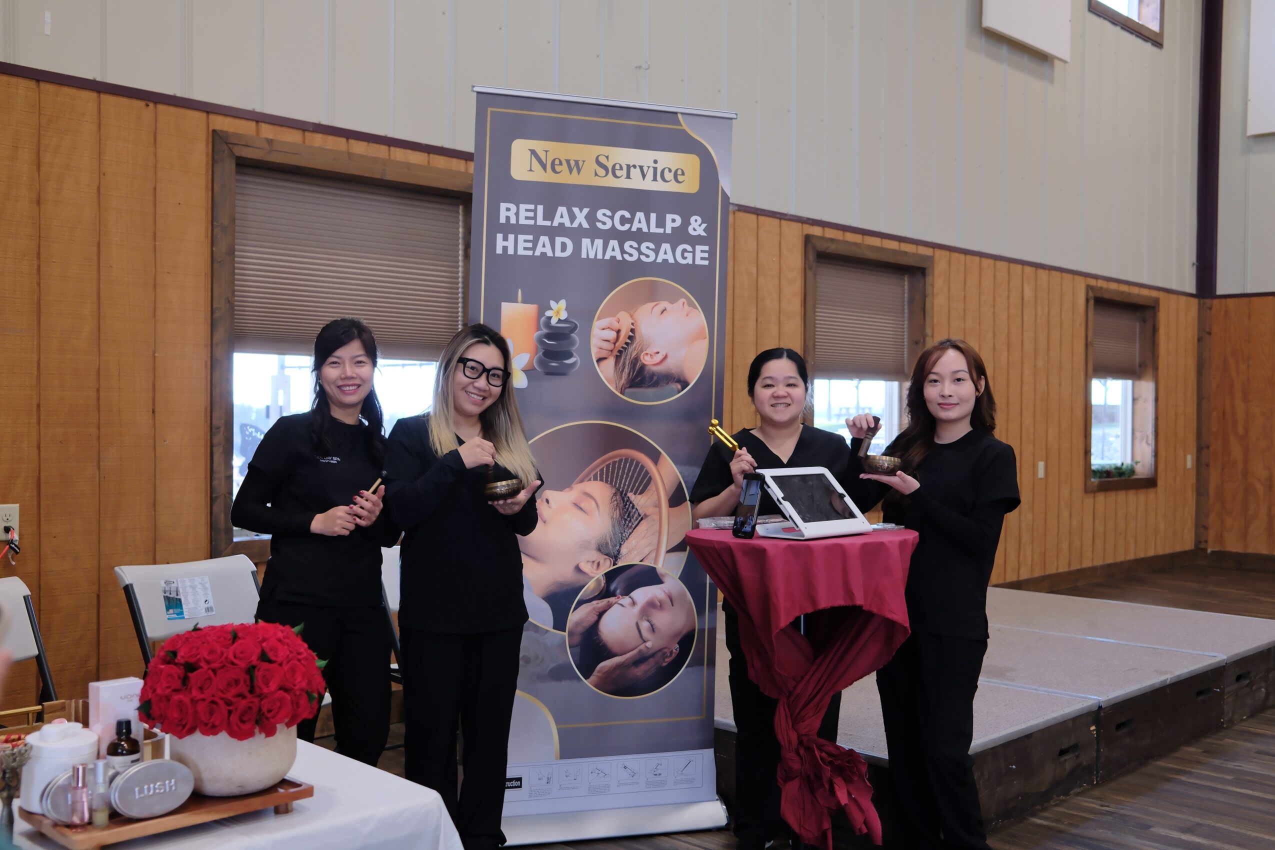 Four women stand next to a tall banner advertising scalp and head massage services, with a table and flowers nearby.