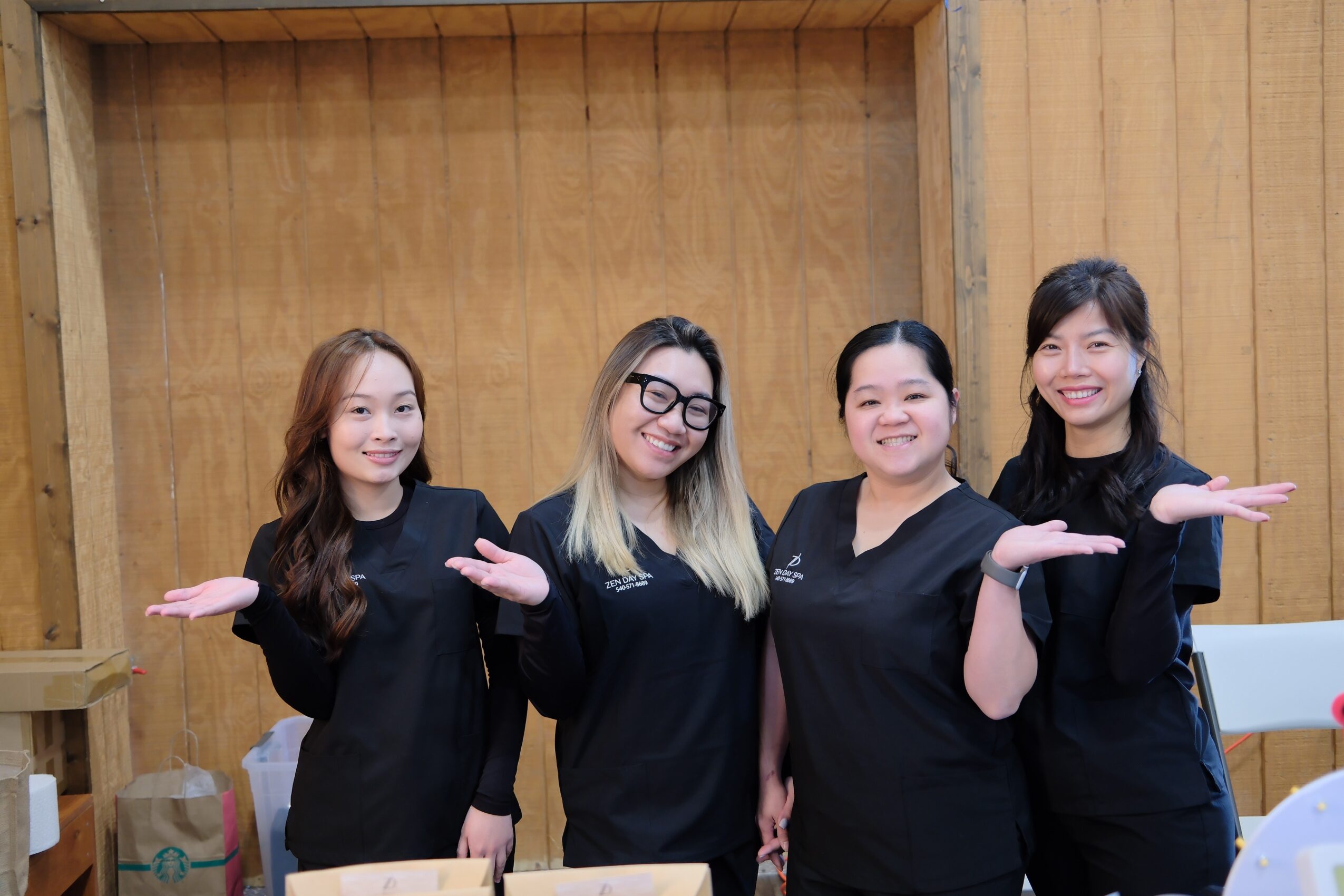 Four women standing indoors, smiling, with wooden wall background, arms extended sideways, wearing black uniforms.