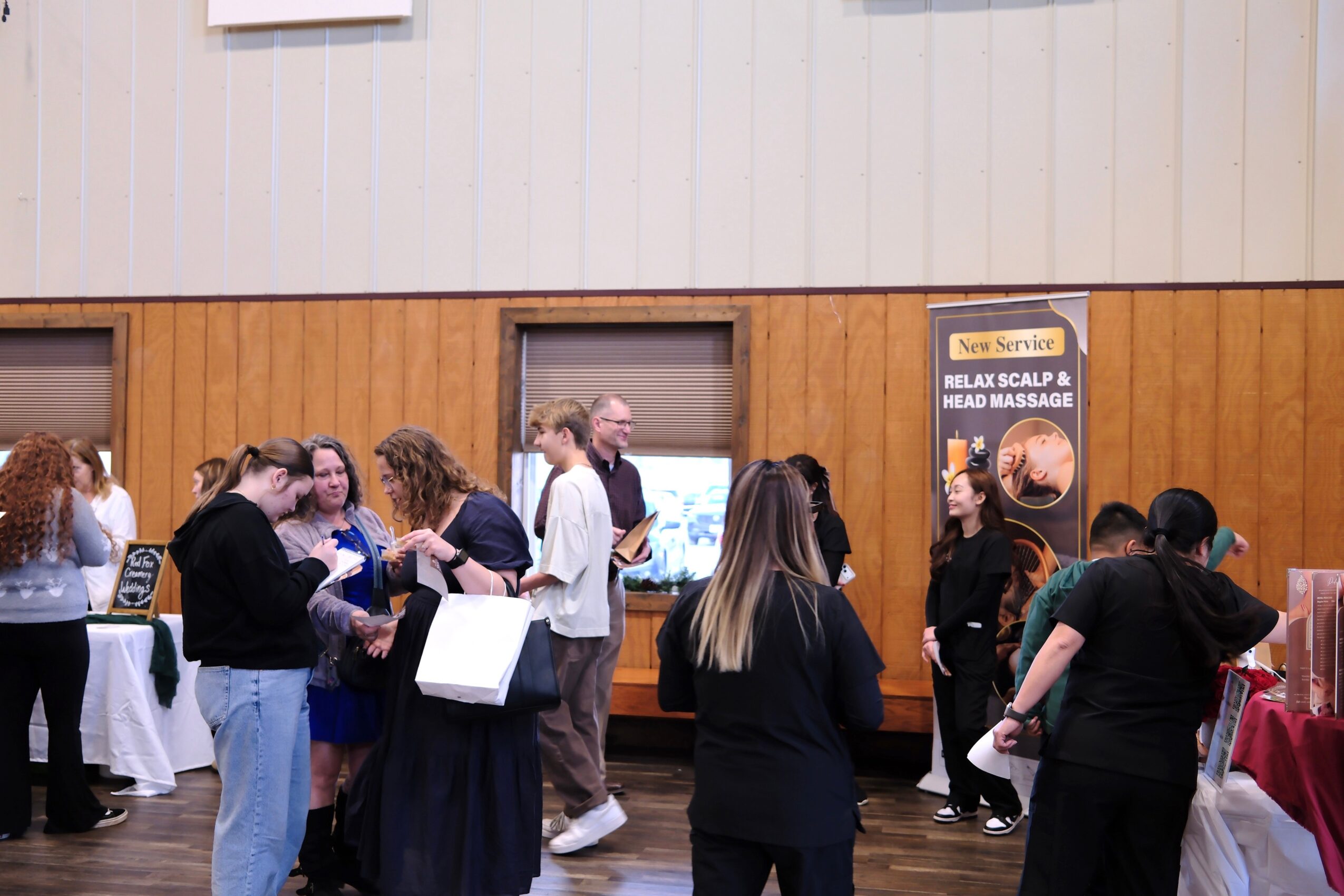 Group of people gathered indoors, some talking and others looking at displays, with a wooden wall and posters in the background.