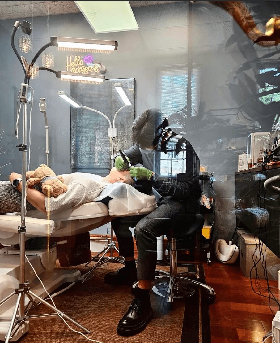 Person sitting in a medical room, with a patient lying on a bed, medical equipment, and a window reflected in the glass.