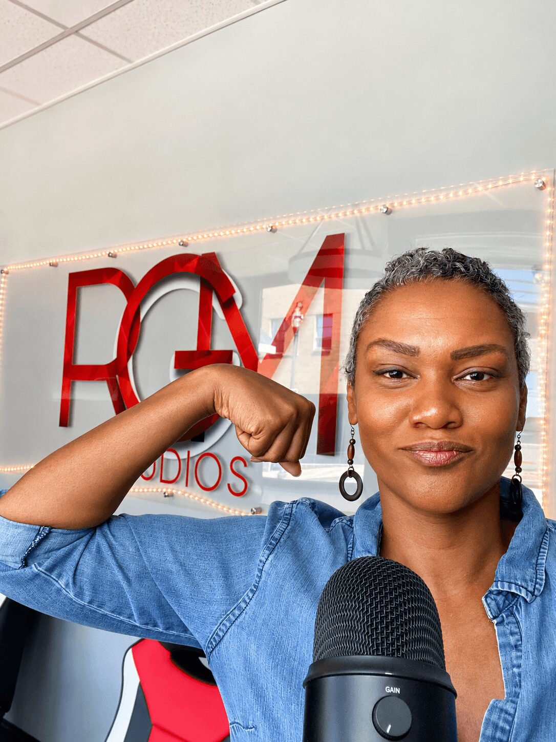 Woman with braided hair and earrings, wearing a denim shirt, flexing her arm in front of a radio station sign.