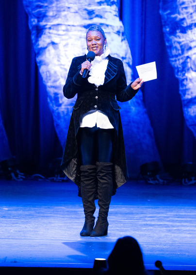 Woman on stage holding a microphone and a paper, wearing a black coat and tall boots, with blue curtains behind her.
