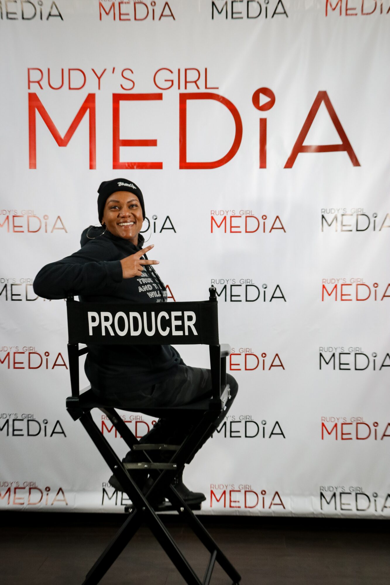 Person sitting on a chair holding a sign that says 'PRODUCER' in front of a backdrop with 'MEDIA' and 'RUDY'S GIRL MEDIA' logos.