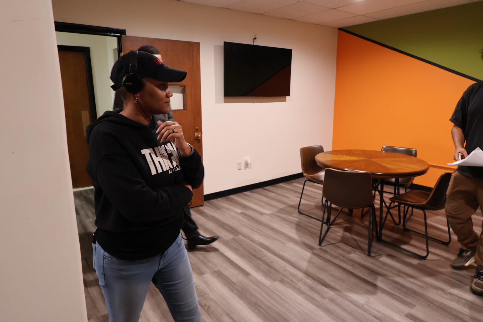 Person wearing headphones and a cap, standing with hand on chin in a room with a table and chairs, wall-mounted TV, and colorful walls.
